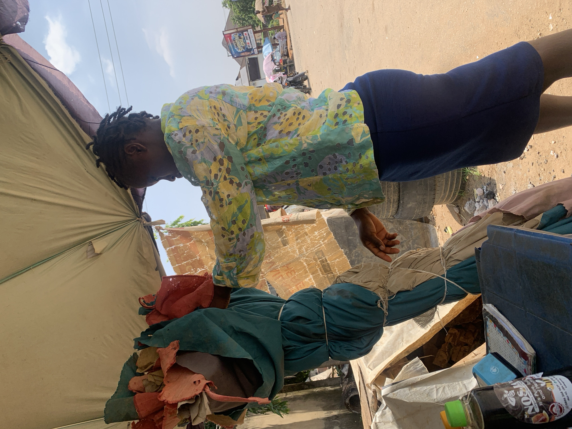 A woman with dark, braided hair stands outdoors under a tan awning, facing a pile of items. She is wearing a yellow and green patterned shirt and a dark blue skirt. Her right hand is extended towards the pile, which is wrapped in teal and beige fabric and tied with twine. To the left of the pile, a bottle of dark liquid with a label featuring a bee and the text "NOSJ..." is visible on a surface with notebooks. The ground is sandy and brown. In the background, there are buildings, other people, a motorcycle, and a billboard with the text "SUPERNATURAL EDGE". The scene appears to be a street market or roadside stall in Abuja, Nigeria.