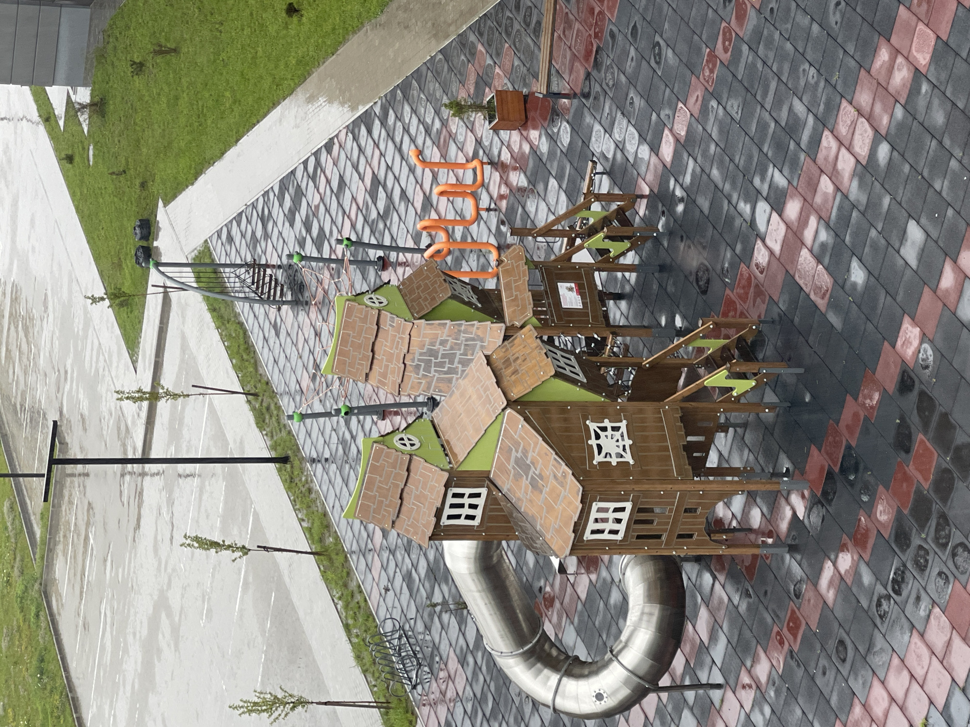 A wet, outdoor playground is shown in Zelenogradsk, Russia. The ground is paved with square tiles in alternating black and red. A large, metal tube slide curves from the left of the frame. A wooden playhouse structure with green roofs and white-framed windows sits in the center. To the right of the playhouse, an orange, winding climbing structure is visible. In the background, a parking lot is wet from rain, with a lamppost and small trees. A grassy area with sparse vegetation and a bench are also present.