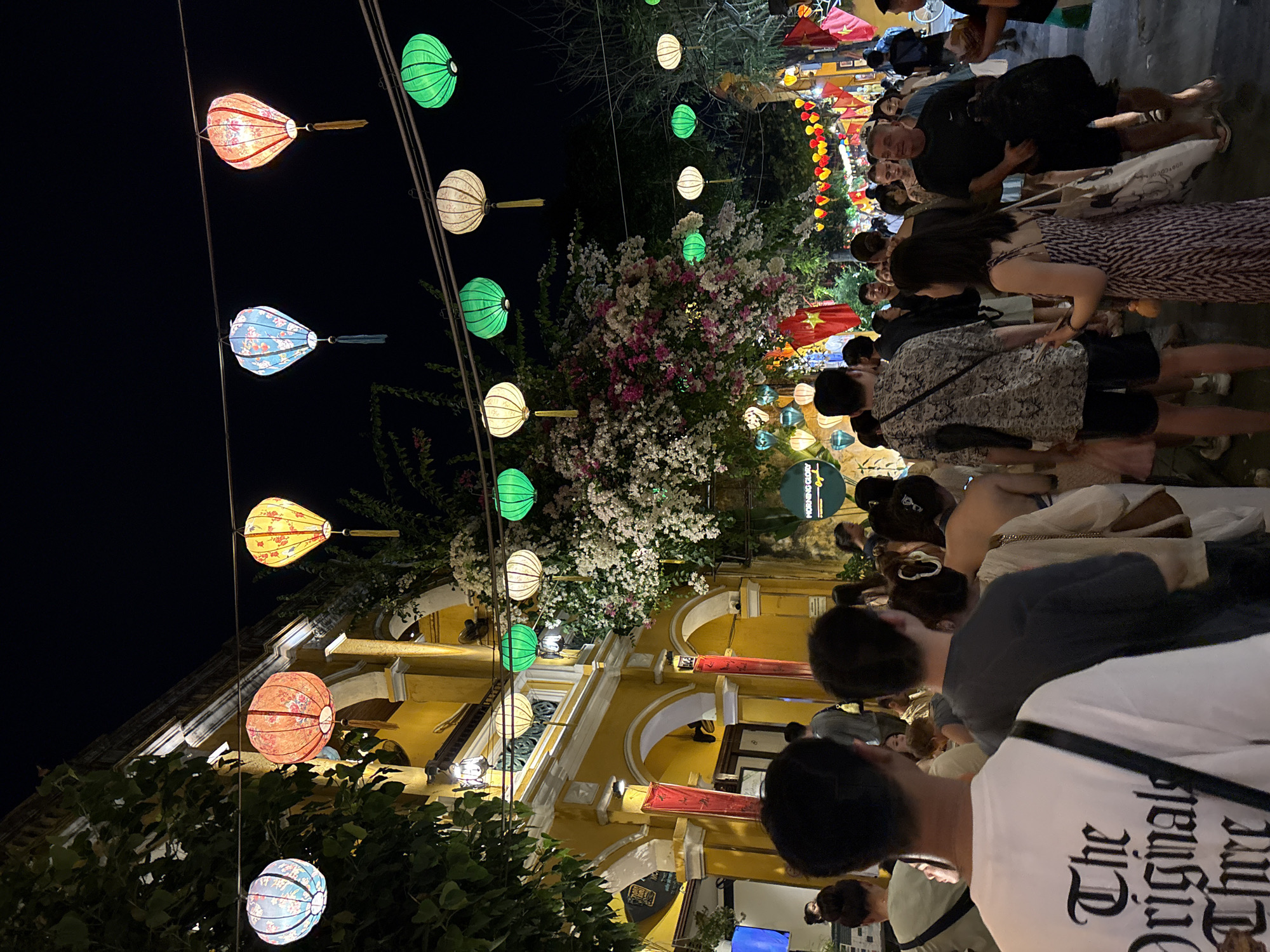 The image shows a street scene in Hội An Ward, Vietnam, at night. Lanterns of various colors and shapes hang overhead, illuminating a crowd of people walking and interacting. The buildings are painted a bright yellow, and some are adorned with flowers. Vietnamese flags are visible in the background. The scene is lively and festive.