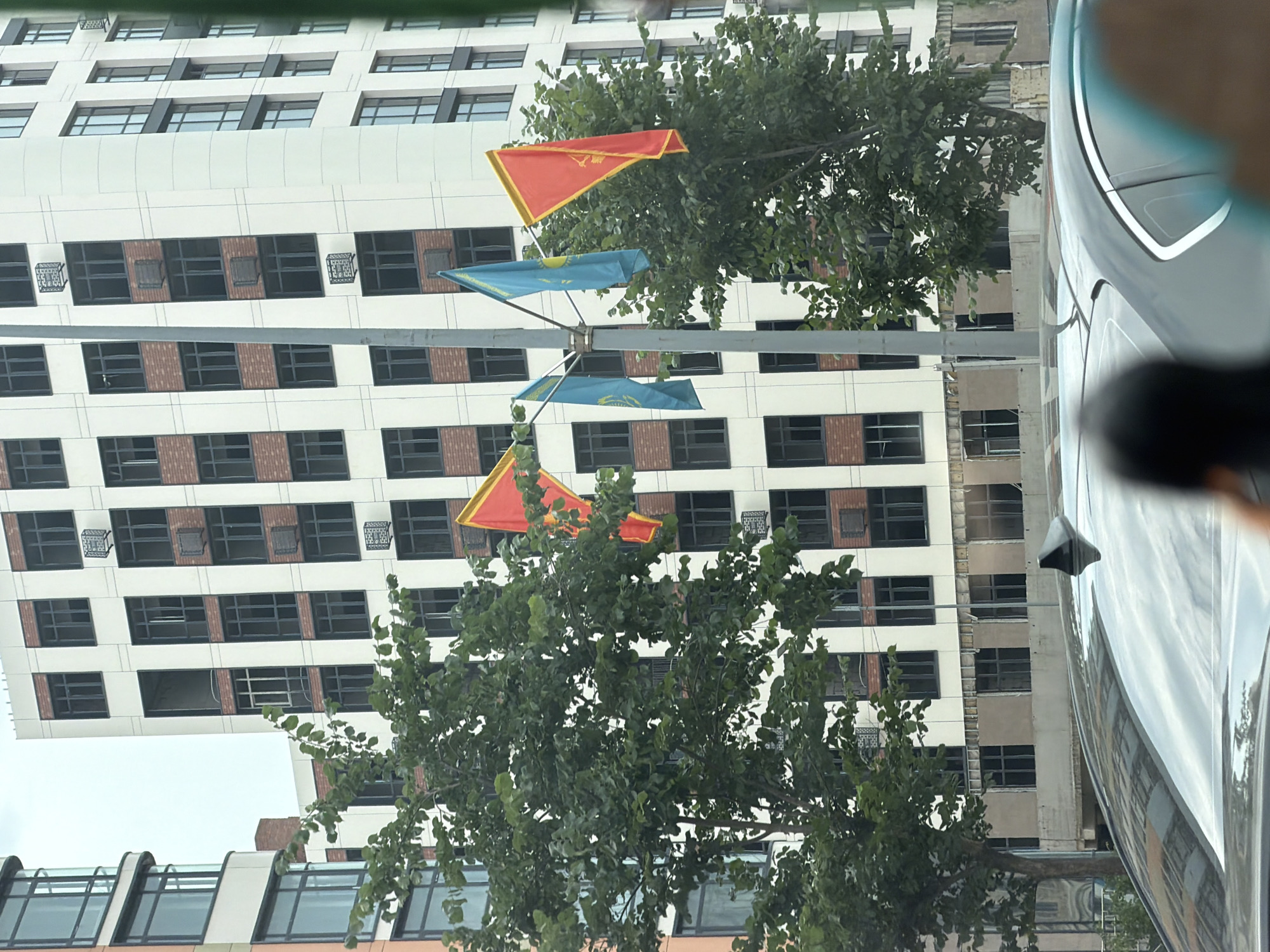 The image depicts a street view in Astana, Kazakhstan, featuring modern architecture and national flags. In the foreground, the roof of a dark-colored car is visible, with a fin-shaped antenna. Behind the car, a tall, multi-story building dominates the scene. The building has a white facade with numerous windows, some of which appear to be black-framed and rectangular. Sections of the building's exterior show exposed brickwork and what appear to be air conditioning units or vents. A street light pole stands vertically in front of the building.
Several flags are displayed on a pole. Two prominent flags are blue with a golden sun and eagle emblem, characteristic of the flag of Kazakhstan. There are also red flags with gold trim, possibly representing regional or celebratory banners. Lush green trees with dense foliage are positioned in front of the building, partially obscuring its lower levels and the flags. The sky above is overcast and light gray. The overall impression is a contemporary urban setting in Astana, likely during daytime.