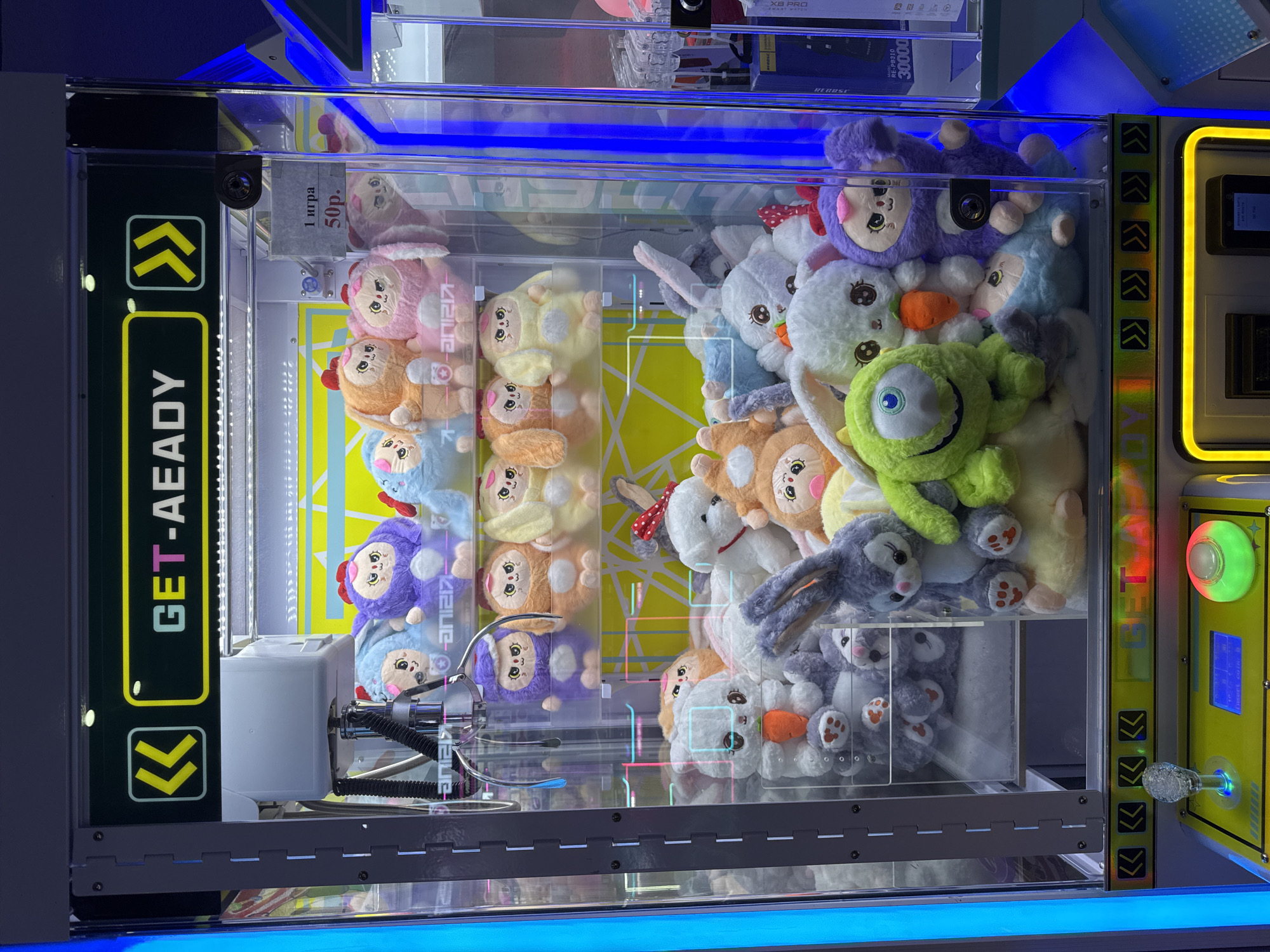 The image shows a claw machine filled with plush toys. The machine has a sign that says "GET-AEADY" and shows prices for playing. The plush toys are various characters, including some that resemble rabbits, and one that looks like the character Mike Wazowski from Monsters, Inc. The machine is illuminated with blue and yellow lights. The background is blurry but appears to be an indoor setting, possibly an arcade or entertainment center. There is no indication of the city of Sochi, Russia, or any specific location within it.