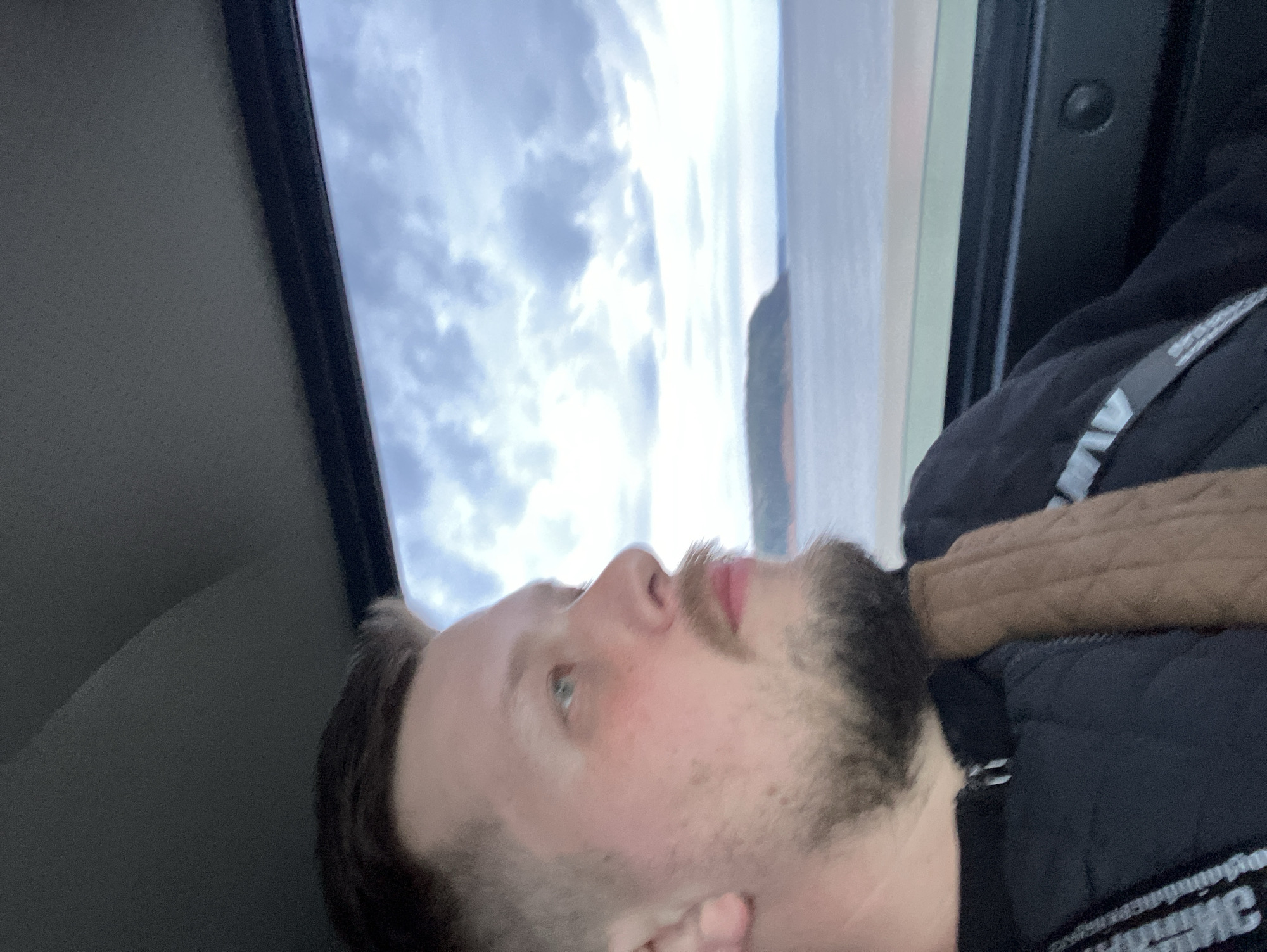 A man with light skin, light brown hair, and a beard is visible from the chest up. He is wearing a black vest with white lettering. The man's face is turned upwards, and the sky is visible through a sunroof. The background suggests a coastal location, possibly Chaikovsky, Russia, due to the presence of a body of water or a wide river.