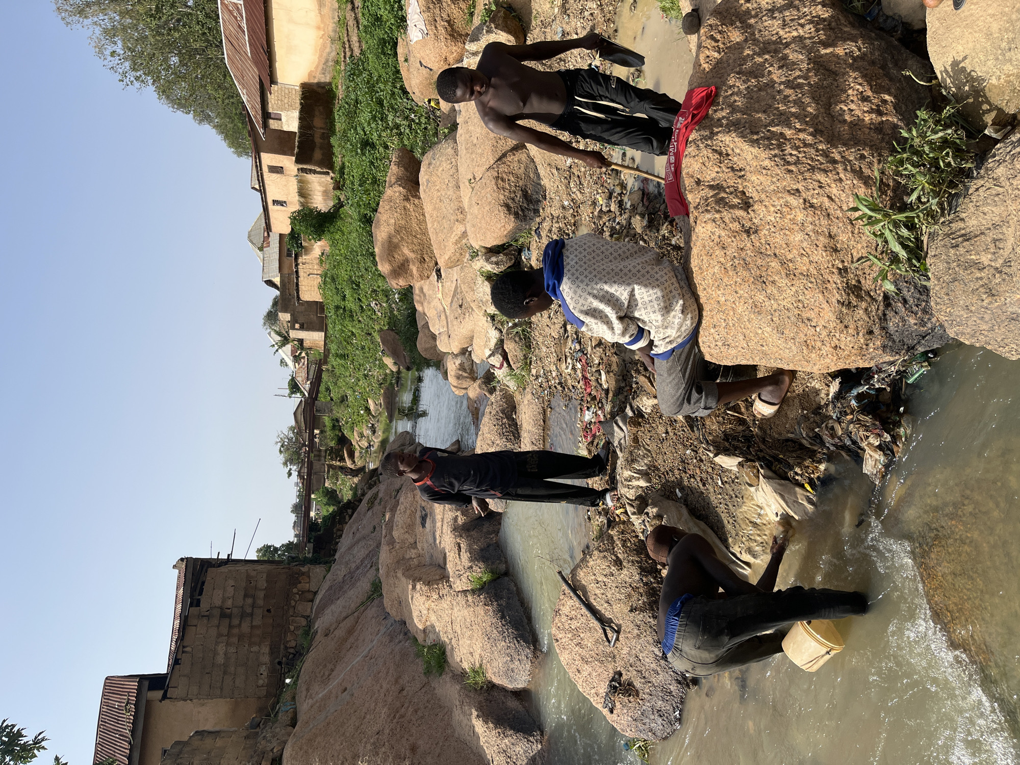 Four young Black males are gathered near a shallow, murky stream in a rocky, uneven terrain. The environment appears to be a semi-urban area with dilapidated buildings and vegetation visible in the background. The stream bed is littered with trash, including plastic bags and debris.
One individual, wearing a black t-shirt with red accents and black pants, stands on the bank, looking down towards the stream. Another individual, dressed in a patterned hooded sweatshirt and grey pants, is seated on a large boulder by the stream. A third person, shirtless and wearing black pants, stands in the water, holding a stick in one hand and a dark object, possibly a shovel head, in the other. The fourth individual, wearing dark pants and a blue waistband, is partially submerged in the stream, holding a yellow bucket and appearing to scoop water or something from the stream bed.
The buildings in the background are made of stone and concrete, with corrugated metal roofs, suggesting a developing or impoverished area. The overall impression is one of a harsh environment where people are interacting with a polluted water source. The specific location in Jos, Nigeria is not identifiable from the visual cues alone.