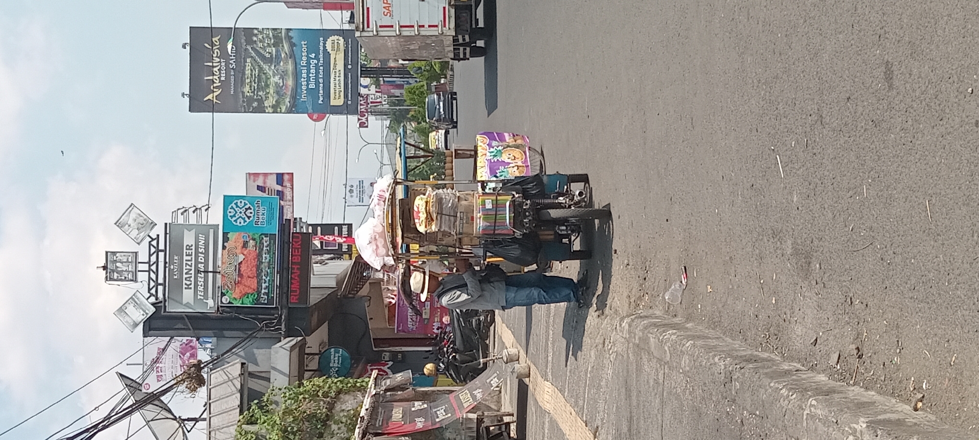 A man wearing a straw hat, a gray long-sleeved shirt, and blue jeans stands on the side of a paved road in Tasikmalaya, Indonesia. He is next to a three-wheeled cart loaded with goods, including packaged items and what appear to be snacks or food items. The cart is attached to a motorcycle, with one wheel visible. The man is looking down and appears to be engaged with something in his hands. In the background, there are buildings with various advertisements and signs, including "KANZLER" and "Andalusia RESORT". Traffic can be seen on the road, with cars and a truck in the distance. The environment is urban, with a sidewalk and street visible. The sky is clear and the lighting suggests a daytime scene.