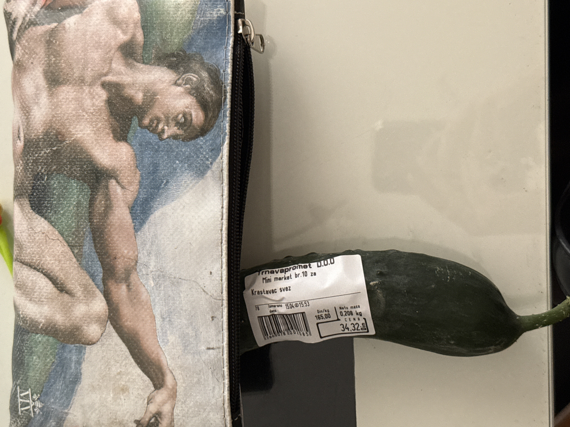 A close-up shot shows a cucumber with a price tag resting on a surface next to a zippered case or bag. The bag features a reproduction of Michelangelo's "Creation of Adam" fresco. The price tag on the cucumber reads "Trnavapromet O.O.O Mini market br. 10 za Krastavac svez," indicating the cucumber was purchased at Trnavapromet Mini Market number 10. The label also shows that the cucumber was weighed on April 15, 2023, at 15:53 and has a net weight of 0.208 kg. The price per kilogram is 165.00 dinars, and the total price is 34.32 dinars. Trnavapromet Mini Market is located in Kragujevac, Serbia.