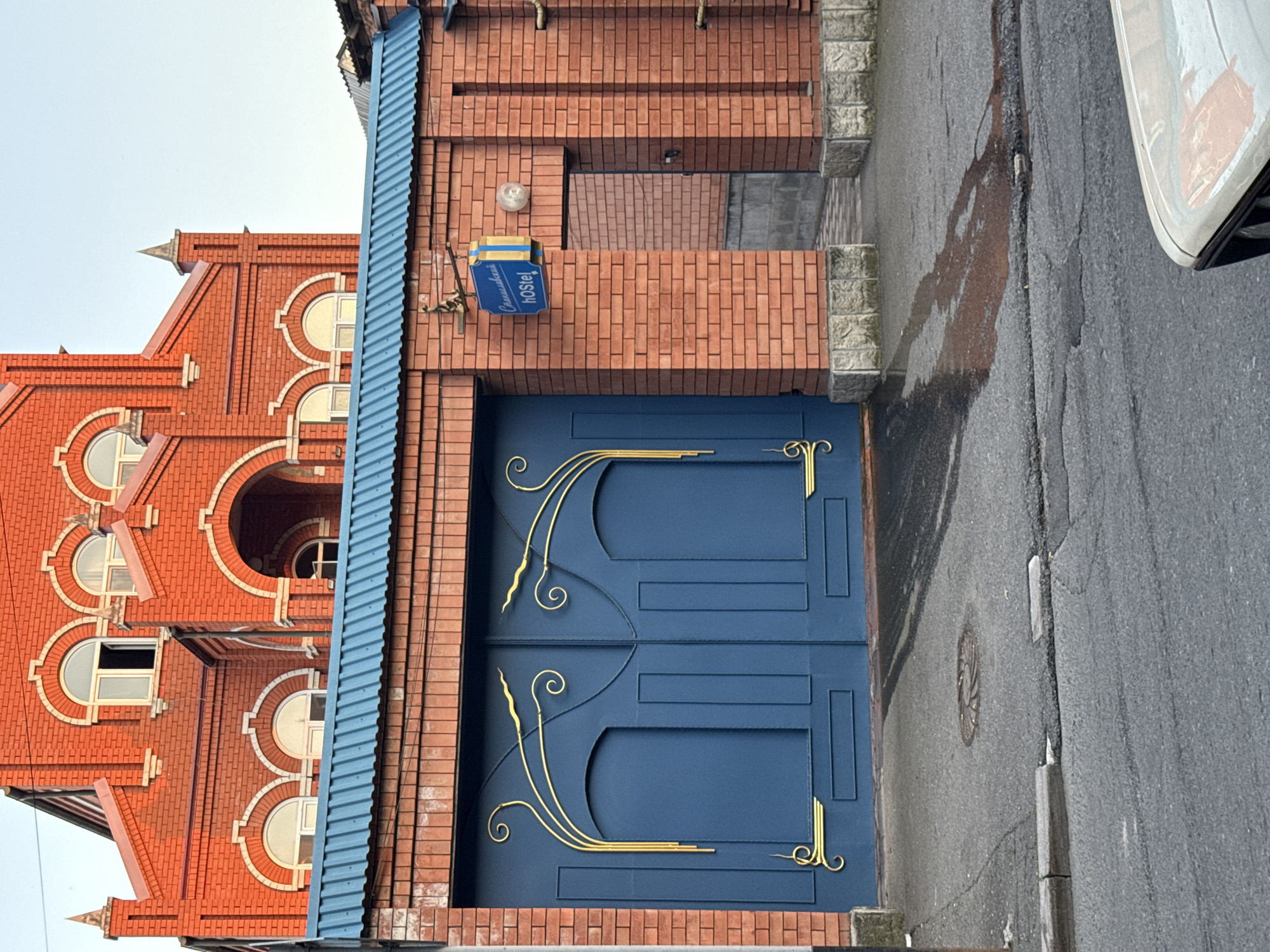 This image shows a large, dark blue metal gate with ornate gold swirling designs, set within a red brick wall. The gate is imposing and appears to be the entrance to a property. Above the gate, the roof of an adjacent structure is visible, made of corrugated blue metal. The brick wall also has a smaller, open doorway to the right of the gate, leading to an interior courtyard or alleyway. A blue sign with yellow lettering is affixed to the brick wall above this doorway, which reads "Славянскaя hOstel".
The architecture of the building visible behind the gate and wall is distinctive, featuring red brick construction with decorative white accents around arched windows and a prominent central archway. The building has a steeply pitched roof with dormer windows. The overall style suggests a potentially older or historically inspired design.
The foreground of the image is a paved road, appearing somewhat worn with cracks and a circular manhole cover visible. There is a patch of wetness on the pavement near the gate, possibly from recent rain or a leak. A portion of a light-colored vehicle is visible in the bottom right corner of the frame. The sky is a pale blue, indicating it is likely daytime. The scene appears to be an exterior urban or semi-urban environment. The location is identified as Sulak, Russia.