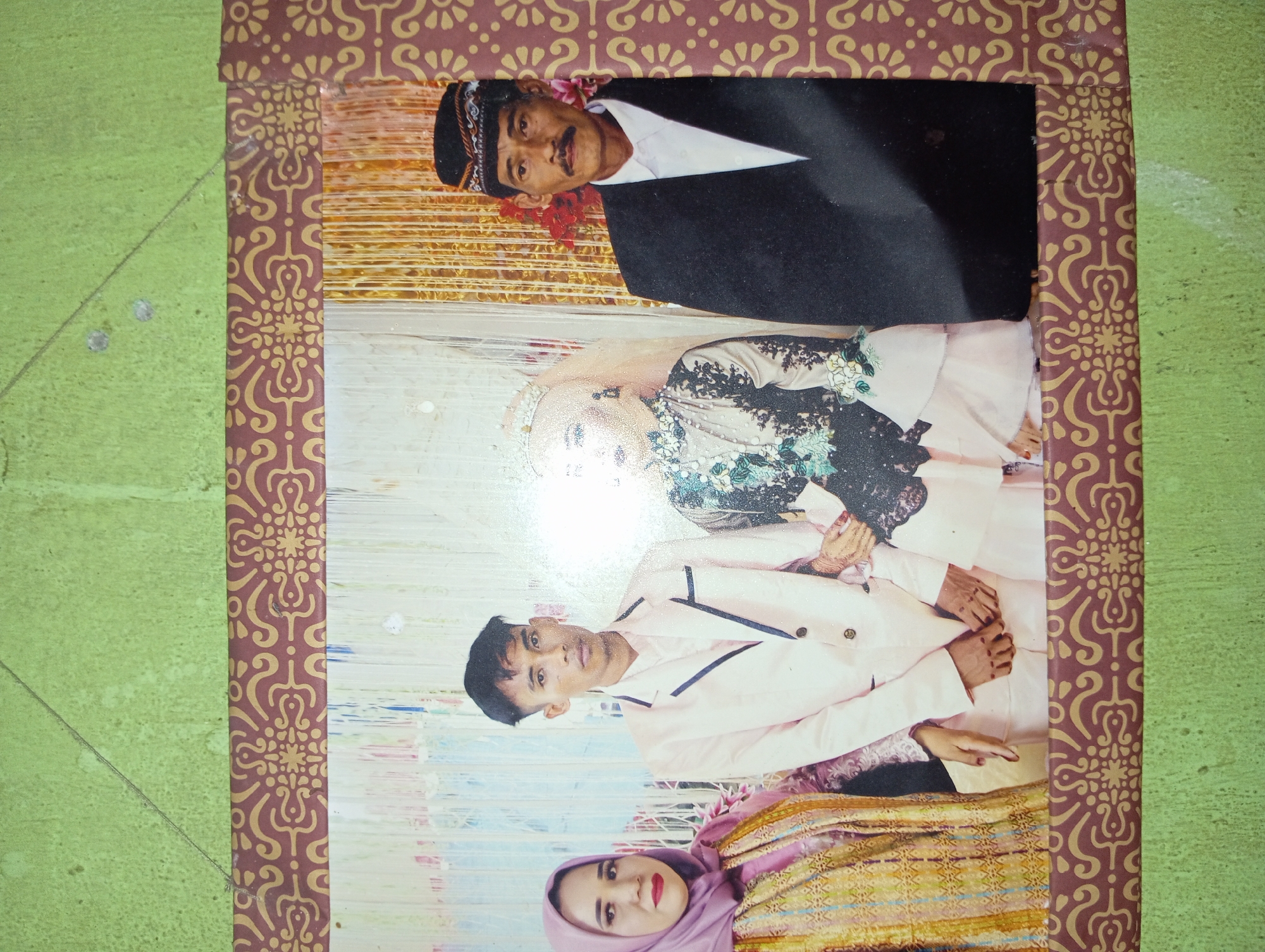 A framed photograph displays four individuals posing for a formal portrait. On the far left, a woman in a light purple hijab and a patterned gold and pink outfit is partially visible. Next to her stands a young man in a light pink suit with a white shirt. To his right is a woman in a white wedding gown and veil, adorned with intricate beading and floral accents. The wedding gown has a light pink underskirt. On the far right, an older man wears a black suit jacket over a white shirt and a black and white patterned cap. His hands are clasped in front of him. The background consists of a curtain-like fabric, with a gold, textured panel behind the older man and to the right of the bride. The photograph is framed by a decorative border featuring a repeating brown and gold pattern, and it is hung on a textured green wall. The setting appears to be indoors, possibly a ceremonial hall or studio in Rejosari, Indonesia, given the attire and context suggestive of a wedding or formal event.