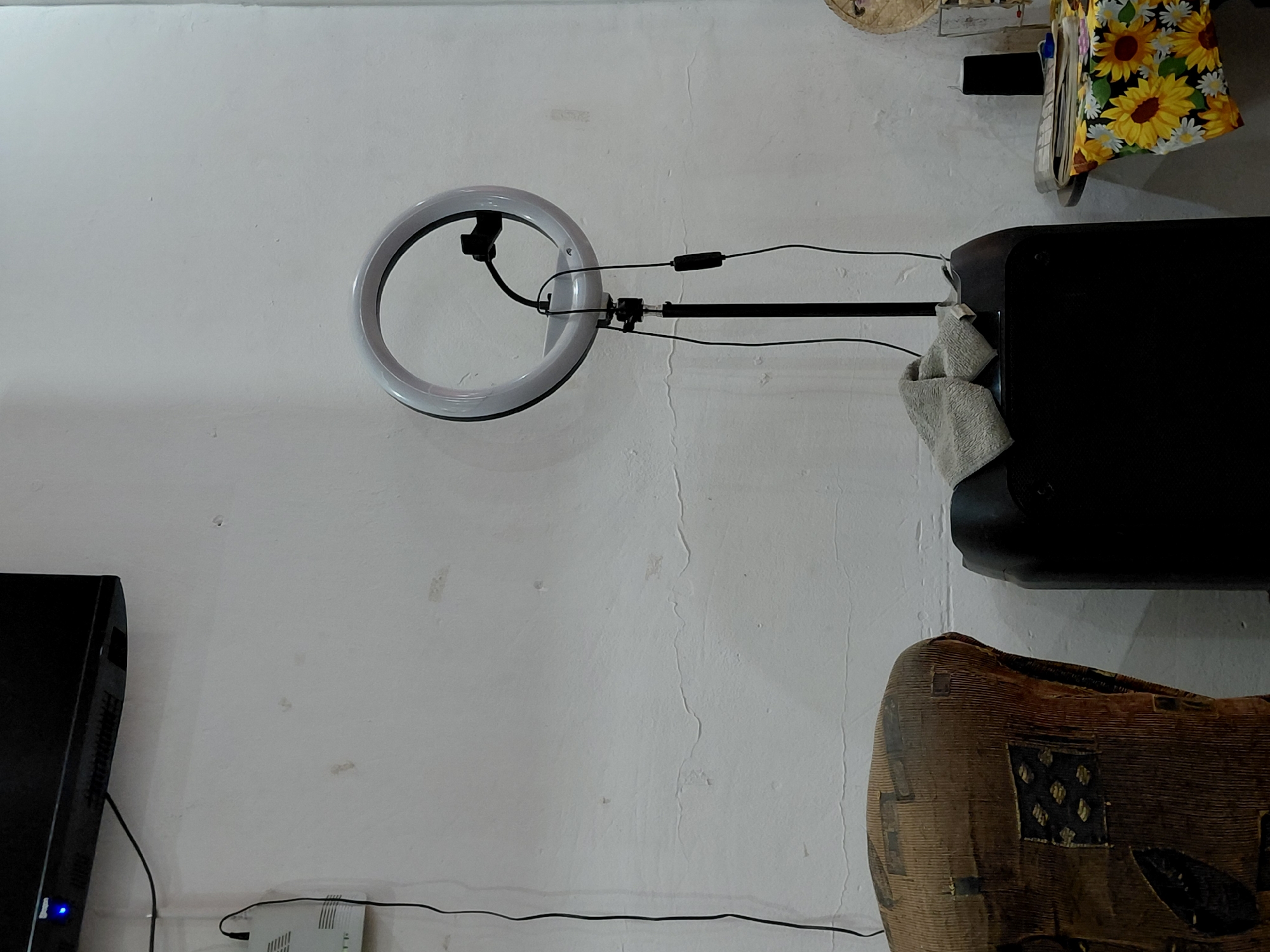 The image captures an indoor scene, likely a corner of a room, displaying various electronic devices and decorative items against a textured white wall. The wall exhibits some wear, including subtle cracks and minor discoloration.
Prominently featured is a ring light, illuminated and mounted on a black stand, positioned in the center-left. Wires from the ring light extend to an inline controller and then off-frame. To the right of the ring light is a large black electronic device, possibly a speaker or sound system, with a light-colored, folded towel draped over its front-right corner. Above this black device, partially visible, is a vibrant yellow fabric adorned with a sunflower and daisy pattern, draped over an unseen object or shelf. A small, woven, straw-like object is also partially visible at the top.
In the bottom-left corner, a black electronic screen, possibly a monitor or television, is visible with a small blue indicator light illuminated. Below it, a white modem or router is connected by black cables that run along the wall. The lower-right corner features a rustic, brown, woven object with geometric patterned patches, resembling a drum or a large decorative basket.
No human subjects are present, and no specific activity is taking place, although the setup with the ring light suggests a space used for video calls, content creation, or similar activities. The scene is illuminated by artificial light, given the glow of the ring light. No visible text is present in the image. The location context provided is Santa Ana, Venezuela.