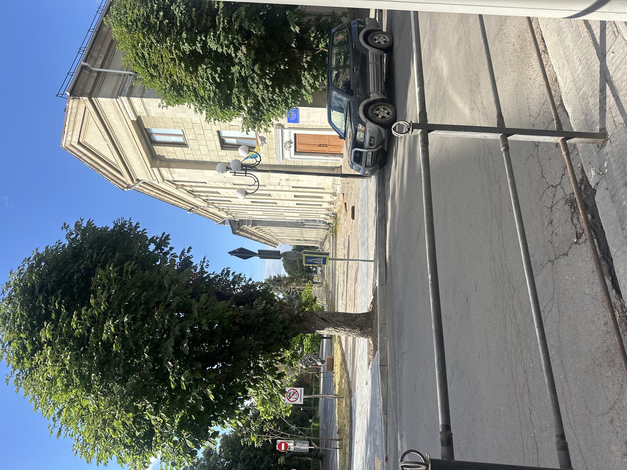 The image captures a sunny day scene in Ternopil, Ukraine, on a street lined with a large building and trees. In the foreground, a black SUV is parked along the curb. The building, constructed of light-colored stone blocks, features classical architectural elements with columns and a triangular pediment. A street lamp with multiple globes stands in front of the building.
To the left of the building and SUV, a large tree with lush green foliage dominates the mid-ground. Several traffic signs are visible on the sidewalk near the tree, including a red circular prohibition sign, a sign prohibiting bicycles, and a pedestrian crossing sign. The sidewalk is paved with grey tiles.
The street itself is asphalt with visible cracks. A metal railing runs along the edge of the sidewalk in the foreground. The background shows more trees and a clear blue sky. The overall impression is of an urban environment with a mix of historical architecture and greenery.