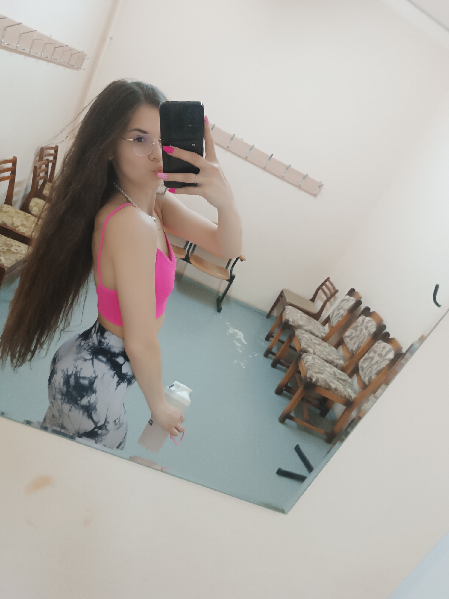 A person with long brown hair is taking a selfie in a mirror. The individual is wearing glasses, a pink top, and grey and white patterned leggings. They are holding a white shaker bottle in their left hand. The environment appears to be a dressing room with chairs lined up against the wall and hooks for hanging clothes. The location is Unknown, Unknown.