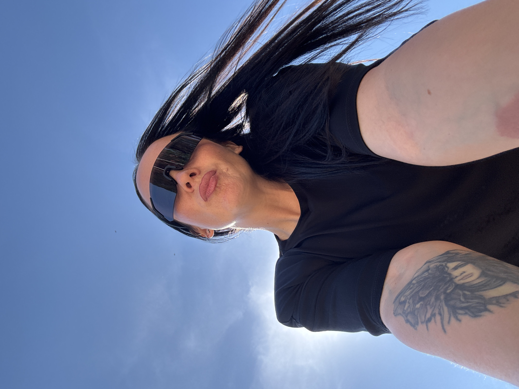 A woman takes a selfie outdoors. The sky is clear and blue, with the sun shining brightly. The woman has long, dark hair that is blowing in the wind. She is wearing sunglasses and a black shirt. Her arm is visible, and a tattoo is present on her shoulder. The image does not provide information about the location in the city of Chaikovsky, Russia.
