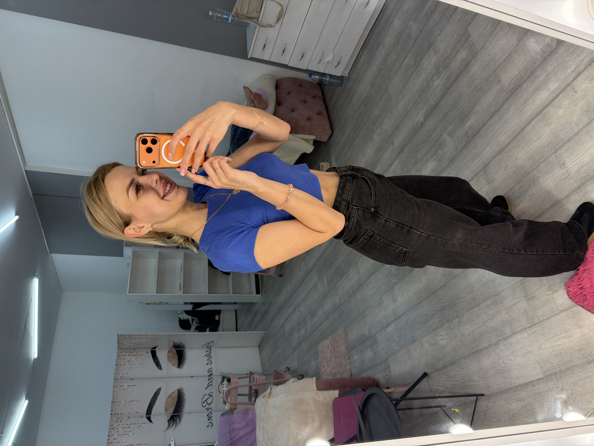 A young woman with blonde hair pulled back in a ponytail is taking a mirror selfie in what appears to be a beauty salon. She is wearing a blue cropped t-shirt and dark wash jeans. She has on several rings and bracelets. The salon has light grey wood-look flooring and bright overhead lighting. In the background, there is a white shelf unit, a pink beauty cart, and a draped screen with an image of eyelashes. A white dresser with drawers is visible on the right side of the frame. A bottle of water and a beige purse are on top of the dresser. The overall impression is a clean, well-lit commercial space.