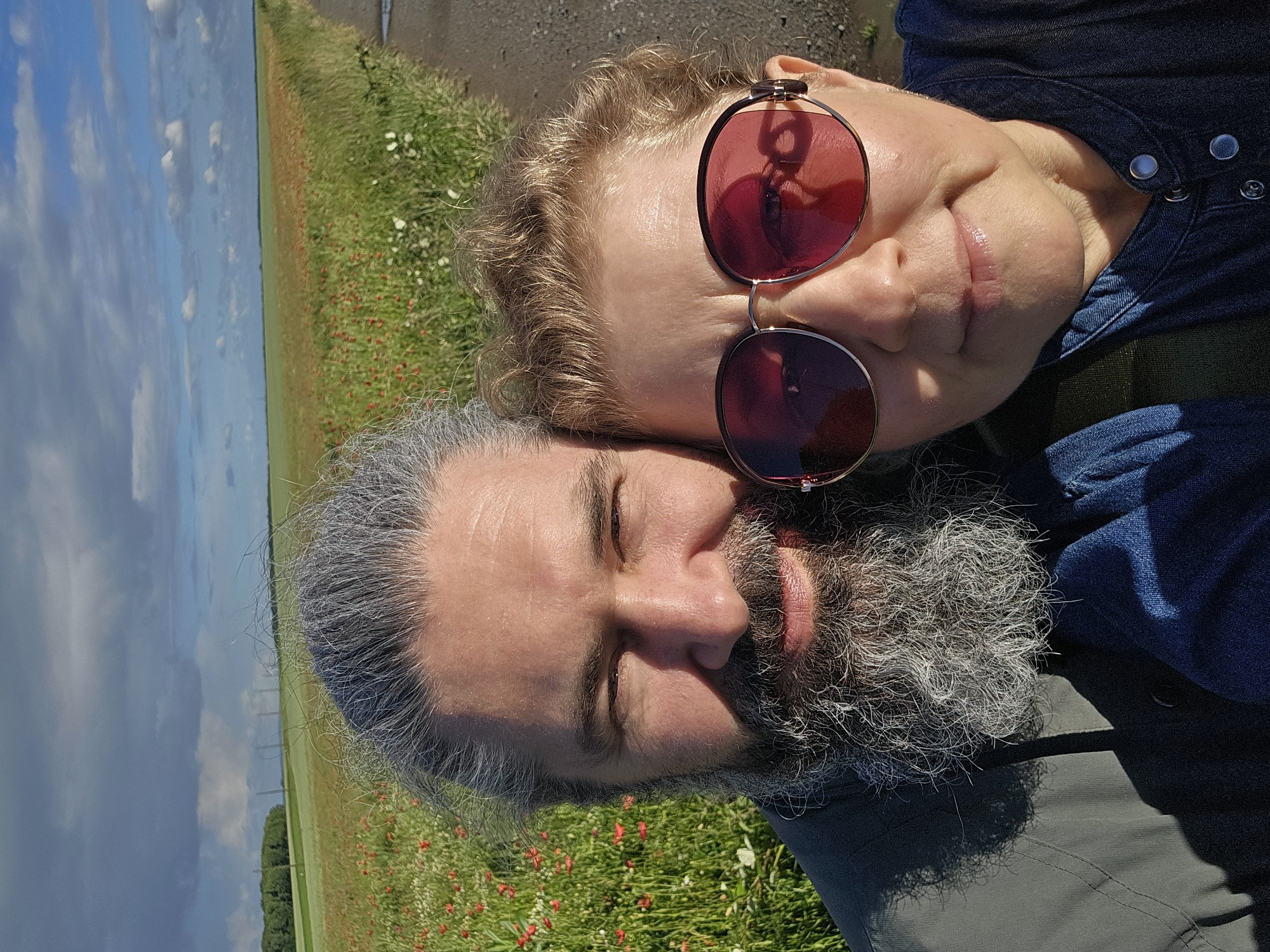 A close-up, selfie-style portrait, originally rotated 90 degrees counter-clockwise, captures two individuals outdoors in Boussu-lez-Walcourt, Belgium. The scene depicts a sunny day with a blue sky and scattered clouds over a vast green field dotted with numerous red poppies and some white wildflowers, bordering a road or path. In the far distance, several utility poles or wind turbines are faintly visible against the sky.
In the foreground, a woman with light, curly hair and striking red-tinted round sunglasses rests her head affectionately against a man with a prominent, full gray beard and graying hair. The woman's face shows a content, subtle smile, with her lips slightly parted. She is wearing a blue denim shirt. The man's eyes are gently closed, conveying a sense of peace or relaxation, and he appears to be wearing a darker jacket or shirt. The bright sunlight casts soft shadows, highlighting facial features and the texture of their hair and clothing. The overall mood is intimate and serene, suggesting a moment of shared enjoyment in a natural, rural setting. There is no visible text in the image.