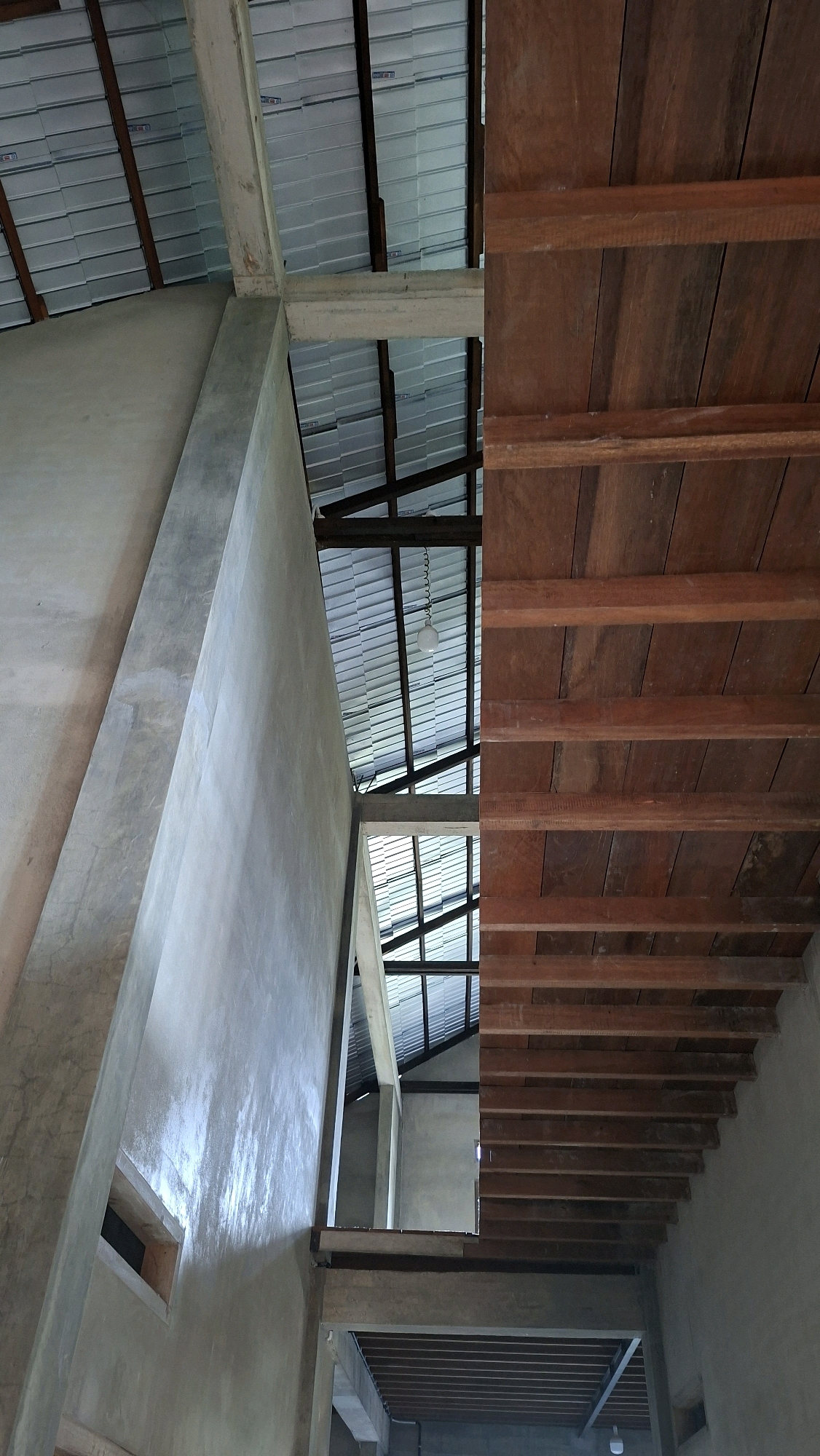 The image depicts the interior of a building under construction, likely a home, in Gronggrong, Indonesia. The scene is divided into two distinct sections by a structural beam. The left side features a raw concrete wall that angles upwards, revealing a portion of a silver, ribbed metal roof structure supported by dark beams. A small, rectangular opening is visible in the concrete wall. The right side of the image shows a ceiling constructed of parallel wooden planks, supported by thinner wooden beams. The lighting is natural, coming from the open roof structure. There are no people or animals visible in the frame. The overall impression is one of unfinished construction, with a mix of concrete and wood elements.
