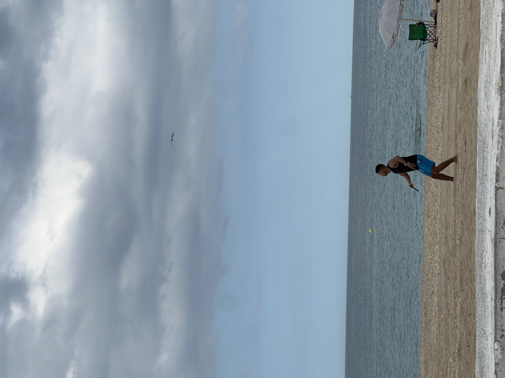 A horizontal shot captures a beach scene in M'Diq, Morocco. On the right, a man in a black tank top and blue shorts is playing tennis near the water's edge. A yellow tennis ball is airborne near him. Further up the beach, a green folding chair sits beneath a striped beach umbrella. In the sky above, a bird flies towards the left, where a cloudy expanse dominates. The sea is calm and occupies the middle ground between the beach and sky. A concrete barrier is visible along the bottom right.