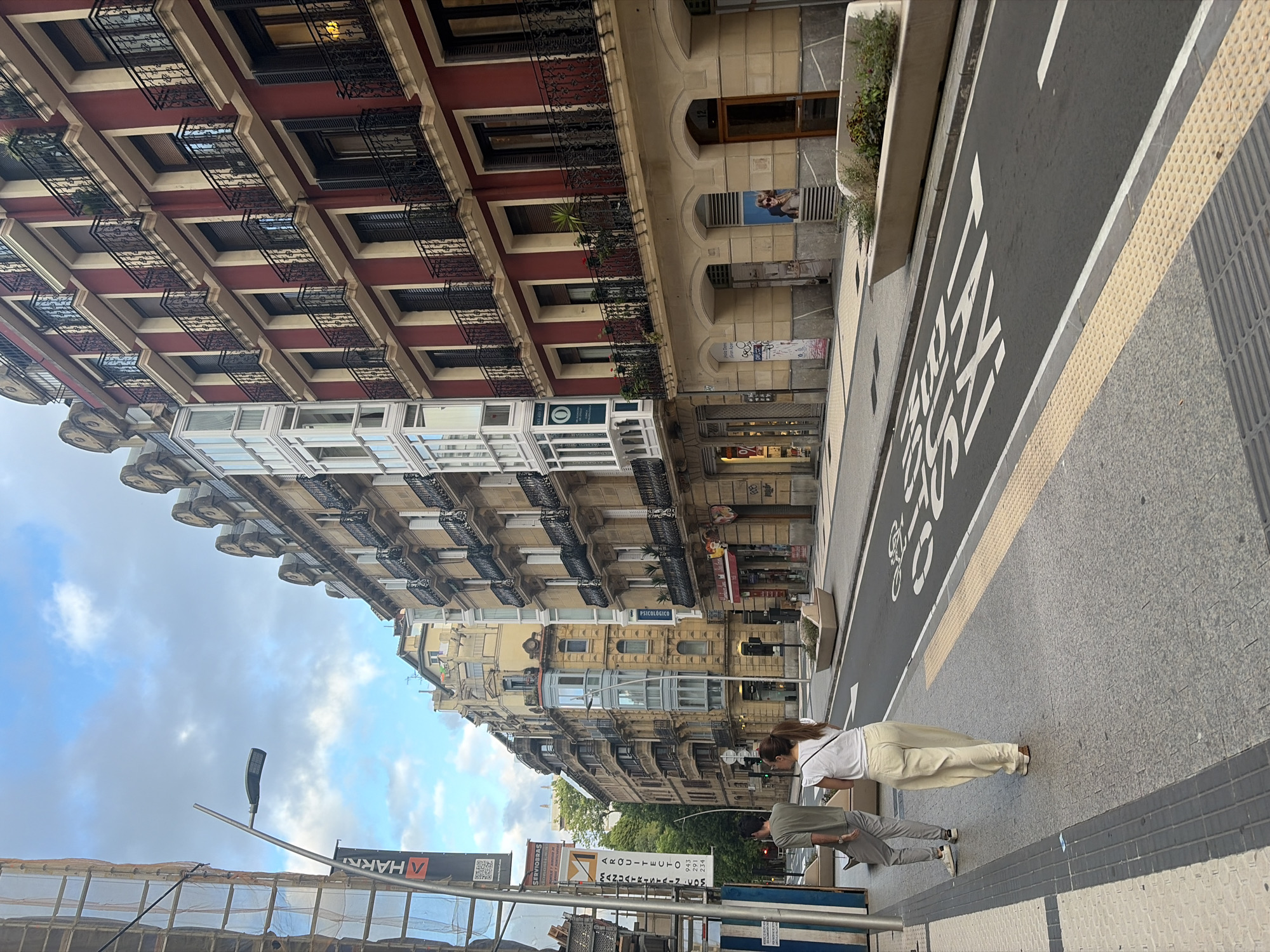 This image captures a street scene in San Sebastián, Spain. The foreground features a sidewalk with two people walking away from the camera. The woman on the right wears a white t-shirt and loose-fitting cream-colored pants, while the man to her left is dressed in a grey t-shirt and grey trousers. They are on a sidewalk next to a street marked with a bicycle lane and the word "TAXI" painted in white letters. The street is lined with multi-story buildings exhibiting traditional European architecture, with some featuring red facades and ornate balconies. Construction scaffolding is visible on the left side of the image, indicating ongoing work on one of the buildings. A streetlamp stands near the scaffolding. The sky is partly cloudy, with patches of blue.