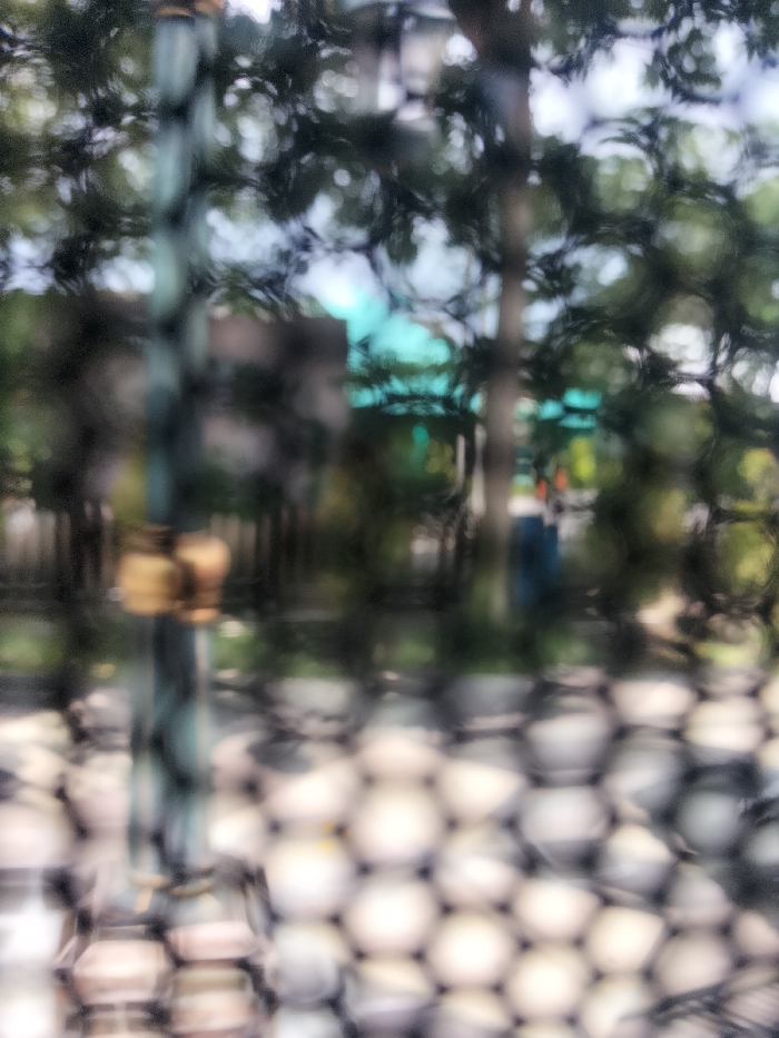 The image shows a blurred view through a wire mesh fence. The location is in Kebomas, Indonesia, at coordinates -7.168 latitude and 112.6 longitude. The scene is partially obscured by the fence, making it difficult to discern details. A large tree in the background appears to be in the foreground, with the fence and other foliage in front of it. There appears to be a green building beyond the tree, which may be a roof. A blurry figure can be seen through the foliage. The image is out of focus and does not provide clear detail about the objects, actions, or interactions in the scene.