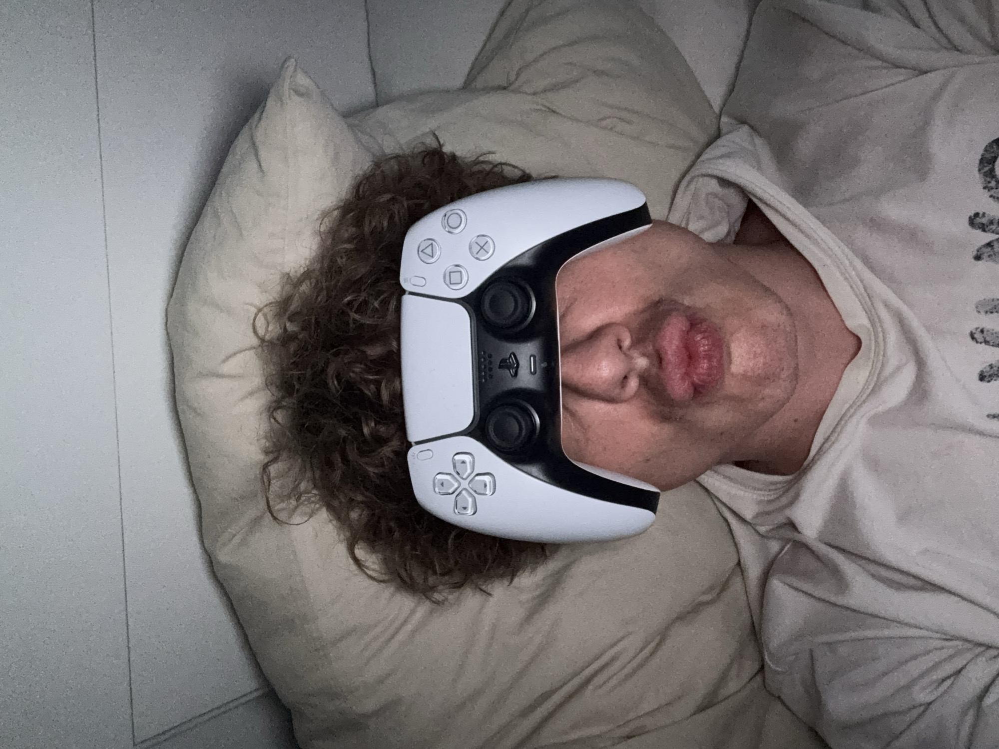 A man is lying on a bed with a white PlayStation 5 DualSense controller resting on his forehead. He has curly brown hair and is wearing a light-colored t-shirt. He is lying on a beige pillow and blanket. The background includes a white wall. The location is not discernible from the image.