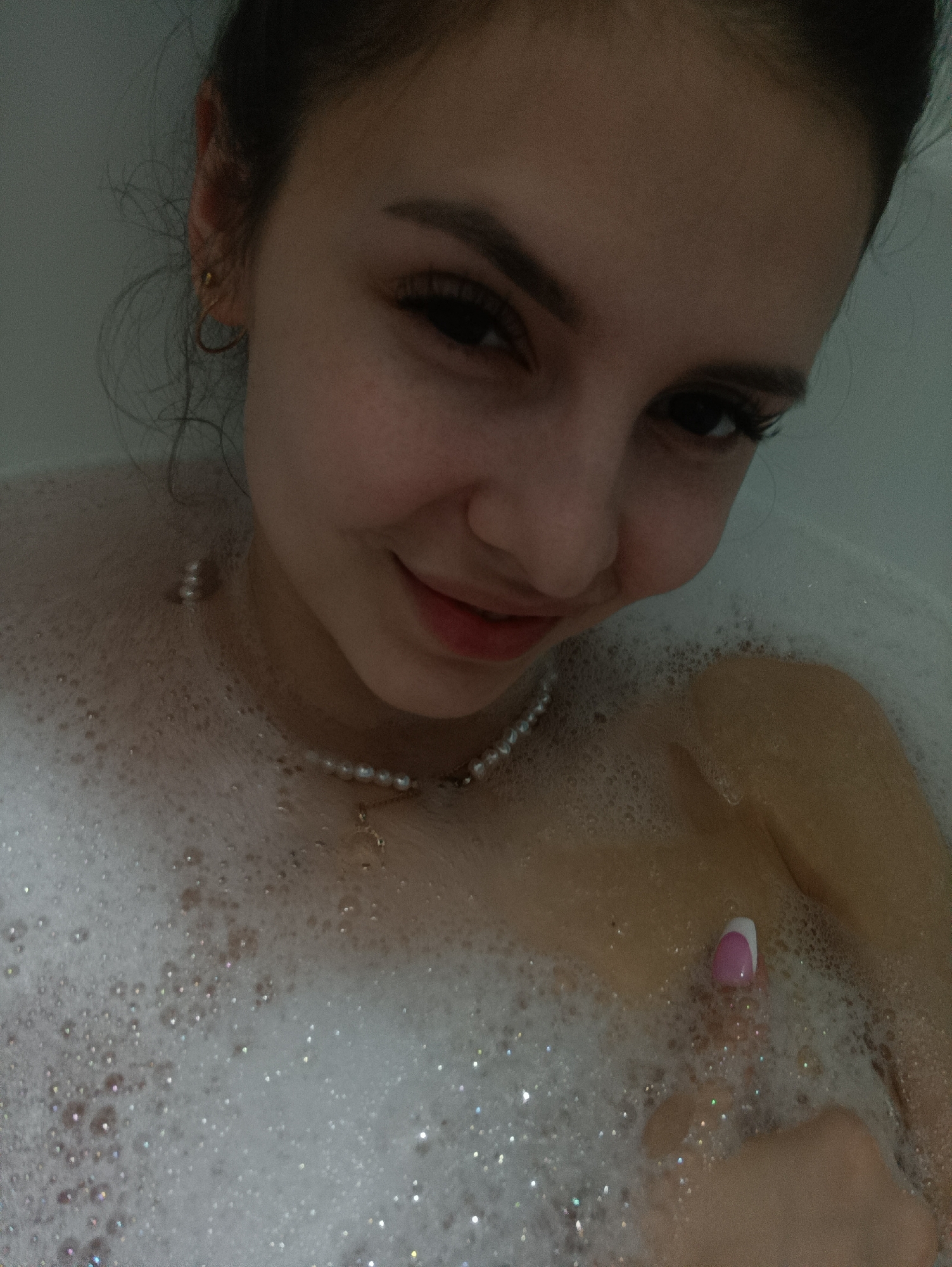 A young woman is photographed in a bathtub filled with bubbles. She is wearing a pearl necklace with a pendant. One of her hands is visible, with a pink and white manicured nail. The location is Unknown, Unknown.