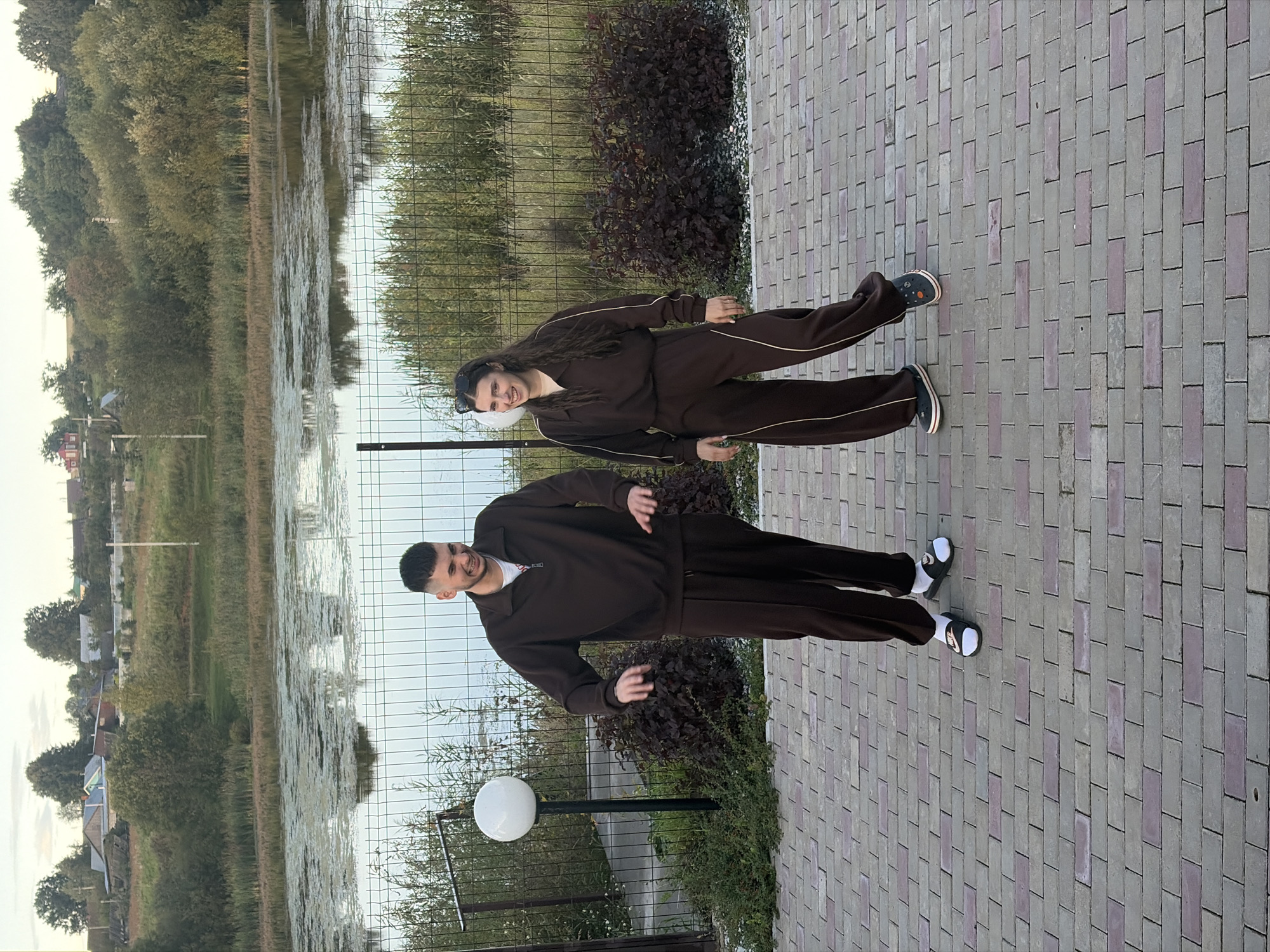 Two individuals, a male and a female, are standing on a paved walkway. The male is on the left, wearing a brown tracksuit and white socks with black slides. He has short, dark hair and is smiling, with his hands held out in a gesturing motion. The female is on the right, also wearing a brown tracksuit, sunglasses on her head, and dark slip-on shoes. She has long, dark, wavy hair and is smiling. Behind them is a body of water with some vegetation on its surface, bordered by a metal fence. Beyond the fence, there is more vegetation and in the distance, buildings and trees are visible under a cloudy sky. The scene appears to be set in a rural or semi-rural outdoor location. The city of Karsha, Russia, is not depicted in this image.