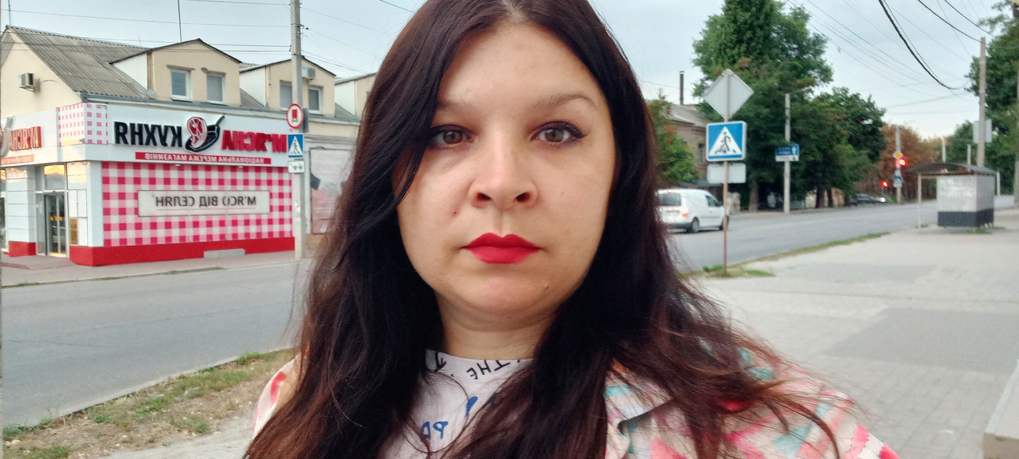 In an urban setting in Kropyvnytskyi, Ukraine, a woman with long, dark hair and bright red lipstick poses for a selfie. She wears a patterned shirt. Behind her, on the left, is a building with a red and white checkered facade, adorned with signs featuring reversed Cyrillic text. One sign prominently displays a red and white checkered pattern. Adjacent to this building, a sidewalk runs along a street. Across the street, a white van is visible, as are trees, traffic lights, and a bus stop shelter. The sky above is overcast.