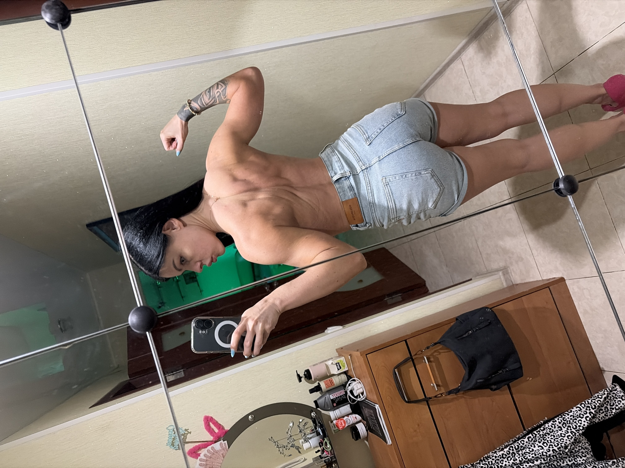 A young woman is posing for a mirror selfie in what appears to be a bedroom or dressing area. She is wearing denim shorts and is shirtless, displaying her back and arm muscles. Her back is to the camera, and she is flexing her right arm, showing off her bicep. She has long, dark hair and tattoos on her right arm.
The environment includes a dresser with various bottles and items on it, a black handbag hanging from a dresser knob, and a leopard-print garment draped over something to the left. The room has light-colored walls with a textured finish. The mirror is large and covers a significant portion of the wall, reflecting the room and the woman. The floor is tiled.
There is no specific indication of the location being in Ufa, Russia, or any other city. The image depicts an indoor, personal space.