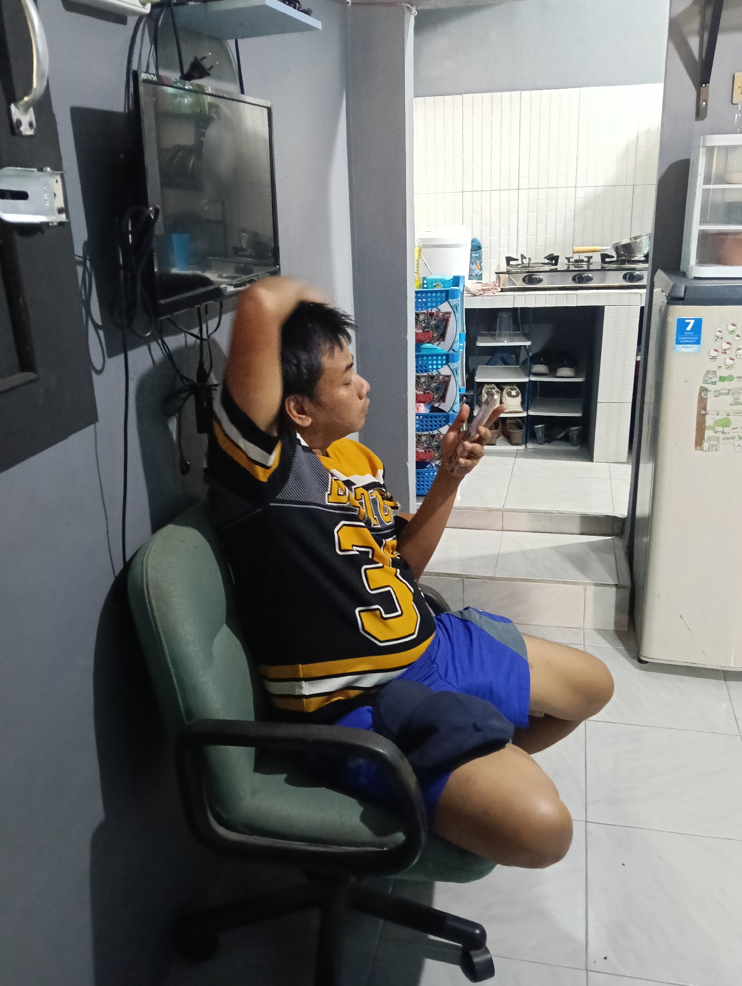 A man sits in a green office chair, his right hand resting on the back of his head, while his left hand holds a mobile phone. He is wearing a black, yellow, and white striped athletic jersey and blue shorts. In the background, a kitchen area is visible with a stovetop, a refrigerator, and shelves filled with various items, including shoes. The scene appears to be indoors, possibly in a residential setting, and the overall lighting suggests it is daytime. The specific location in Tasikmalaya, Indonesia, cannot be determined from the visual information provided.