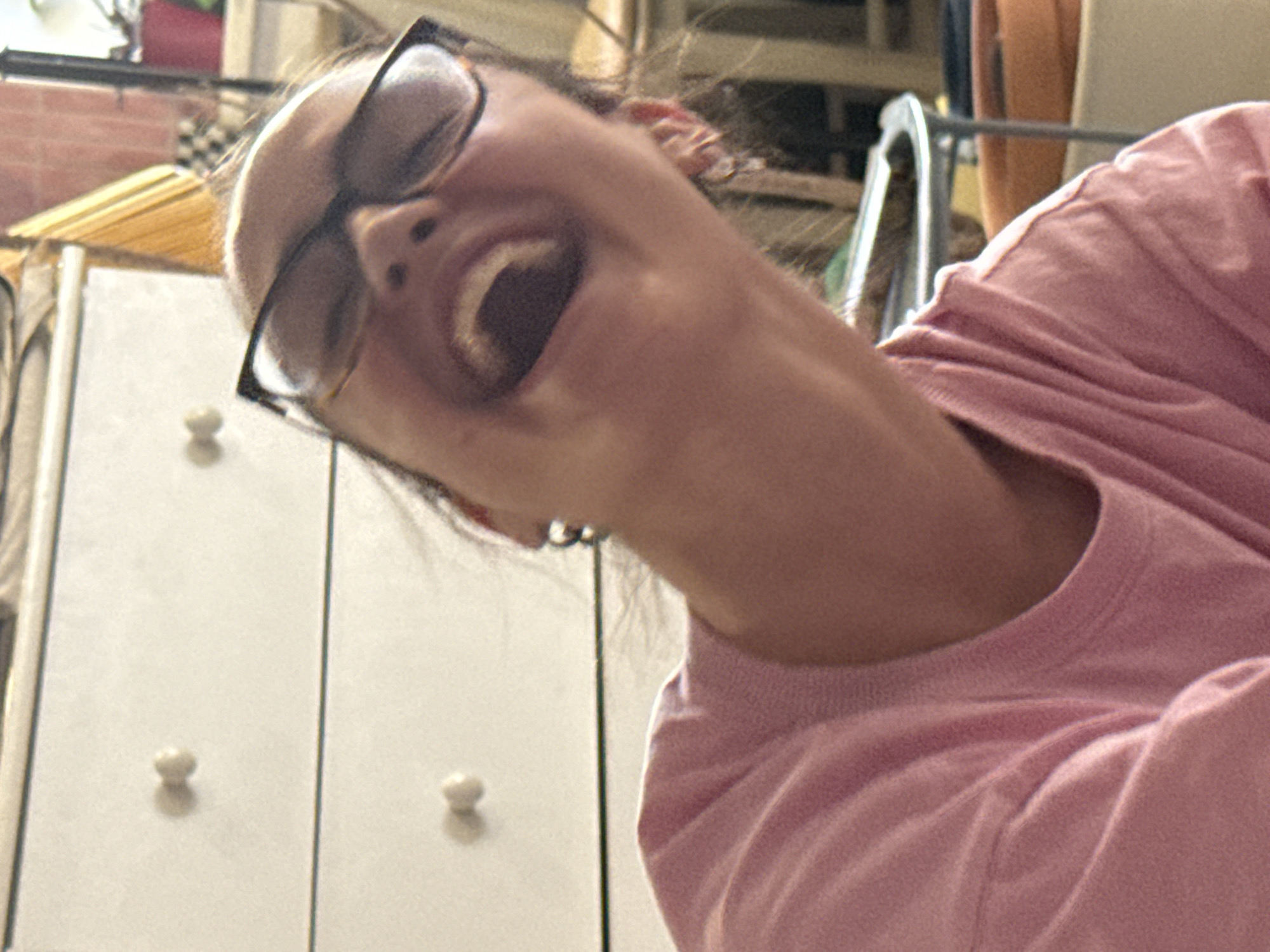 A person with glasses and a pink shirt is laughing with their head tilted back. Behind the person is a white cabinet with three round knobs. The location is not identifiable as Krasnoyarsk, Russia.