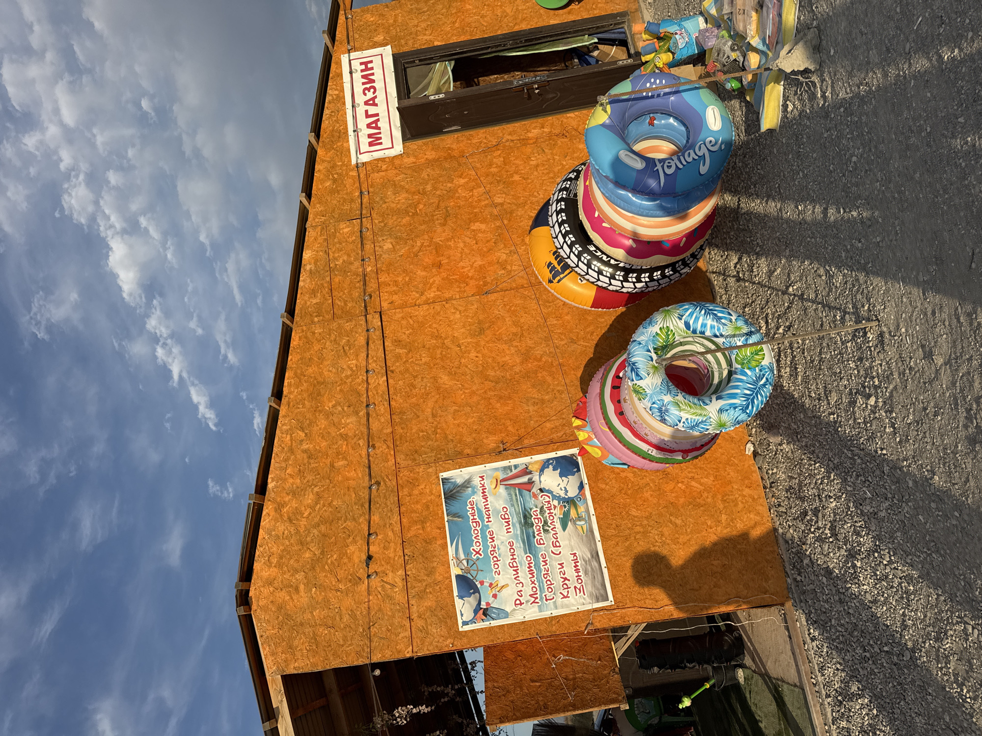 The image depicts a small, open-air shop with an orange plywood facade, located on a gravel surface, likely in Zharbulak, Kazakhstan. The shop has a sign that reads "MAGAZIN" in red lettering, indicating it is a store. In front of the shop, several inflatable swimming rings are stacked, some with colorful patterns. A sign on the wall lists items for sale, including "Cold drinks," "Hot snacks," "Draft beer," "Mojito," "Hot food," "Rings (balloons)," and "Umbrellas." To the left of the shop, a shadow of a person is cast on the wall. The sky above is blue with scattered clouds. The overall scene suggests a beach or recreational area where beach supplies and refreshments are sold.