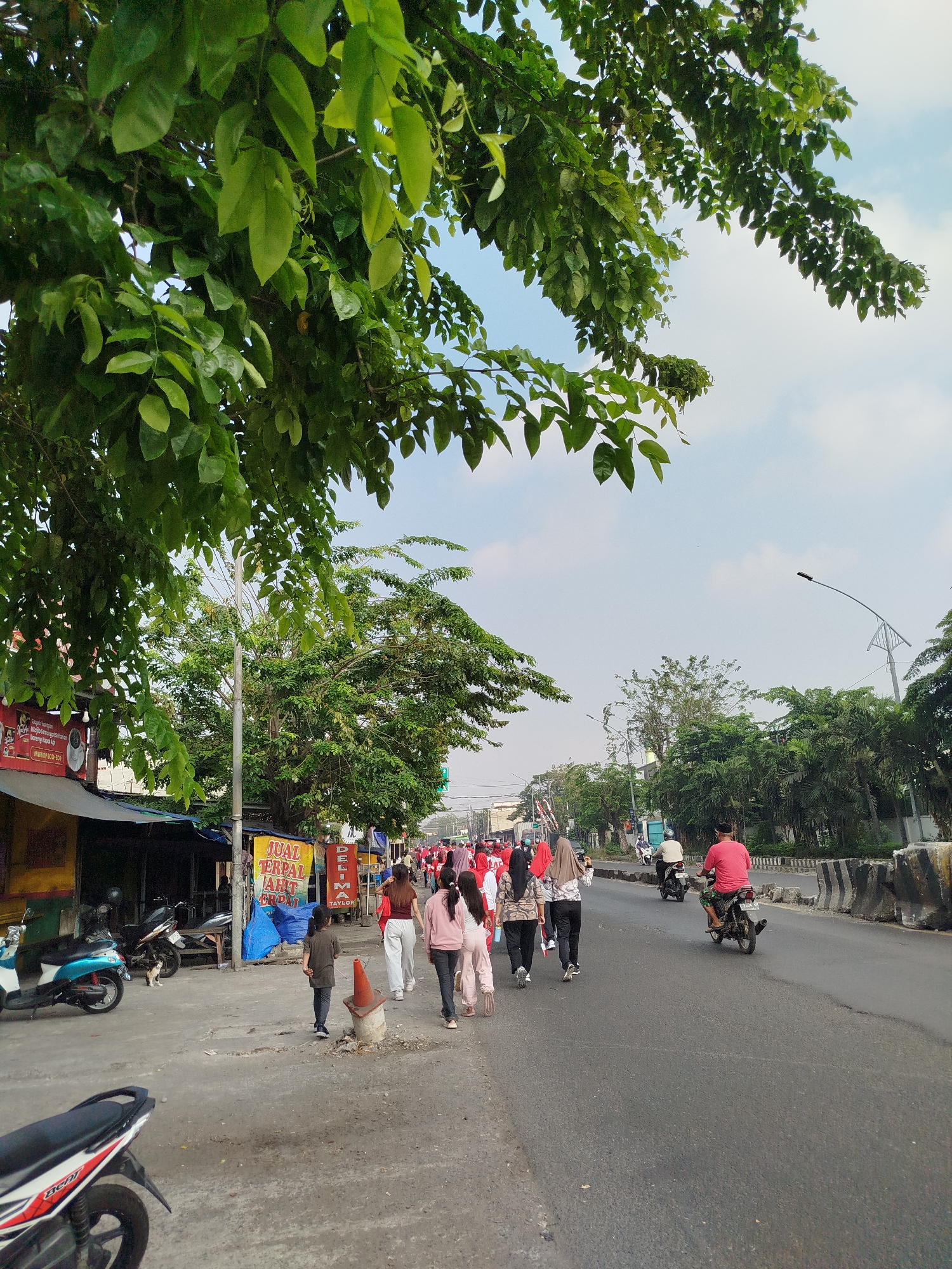 The image depicts a street scene in Surabaya, Indonesia, likely during a daytime event. On the left, there are small roadside stalls with awnings and various merchandise displayed, including a large blue tarp and signs with Indonesian text. Several motorcycles are parked in front of the stalls, including a white and blue scooter in the foreground and a black motorcycle further back. A small cat can be seen near the black motorcycle.
A group of people, mostly women wearing headscarves and casual clothing in various colors, are walking along the sidewalk and into the street. Some are holding small Indonesian flags. A young boy in a grey shirt is walking away from the viewer on the sidewalk, near a traffic cone.
The street is paved and has at least two lanes. A man in a red shirt is riding a motorcycle in the right lane, moving away from the viewer. Another person on a motorcycle, wearing a light-colored shirt, is also in the right lane ahead of the man in red. Concrete barriers line the right side of the road, and behind them, lush green trees and palm trees are visible. Overhead, the sky is partly cloudy. The branches of a large leafy tree extend into the top left of the frame, casting shadows on the scene. The overall impression is of a bustling local event or procession in an urban Indonesian setting.