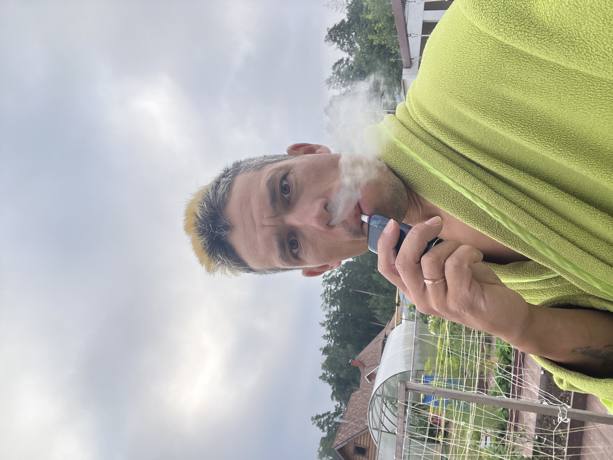 A man with dyed blonde hair is holding a vaping device to his mouth and exhaling a cloud of vapor. He is wearing a lime green bathrobe. The man is outdoors, with a rural landscape behind him. There are several houses and greenhouses visible, along with trees. The sky is cloudy. The man appears to be in a suburban or rural area, possibly on his property, given the presence of greenhouses and a residential structure. The location is Chaikovsky, Russia.