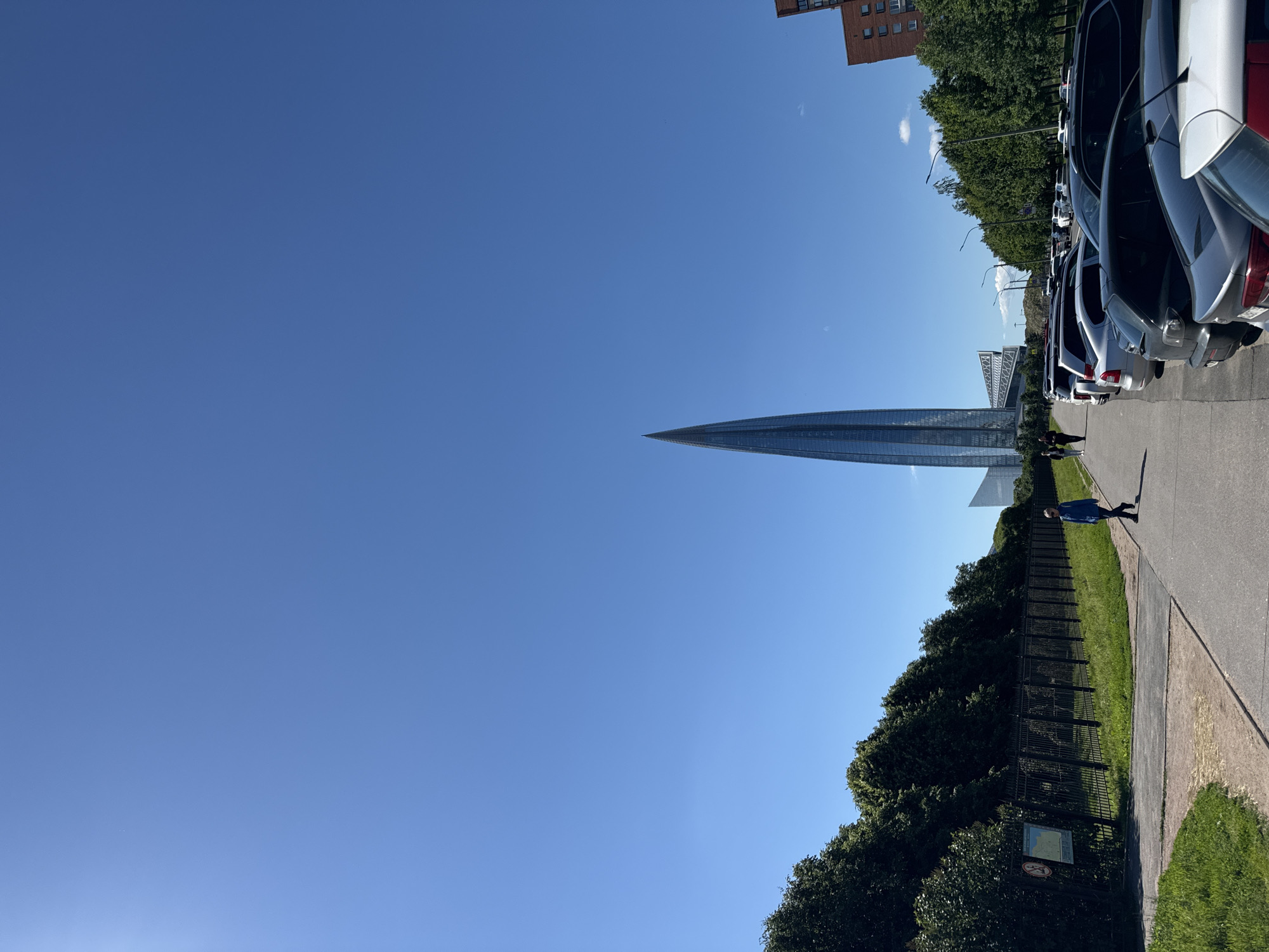 The image depicts a clear, sunny day in Saint Petersburg, Russia, with a prominent skyscraper dominating the skyline. The skyscraper, which appears to be the Lakhta Center, has a distinctive spiral design and is made of glass. In the foreground, a row of parked cars lines a street next to a pathway. A person wearing a blue jacket is walking on the pathway. A metal fence with greenery behind it runs along the side of the pathway. In the distance, other buildings and trees are visible.