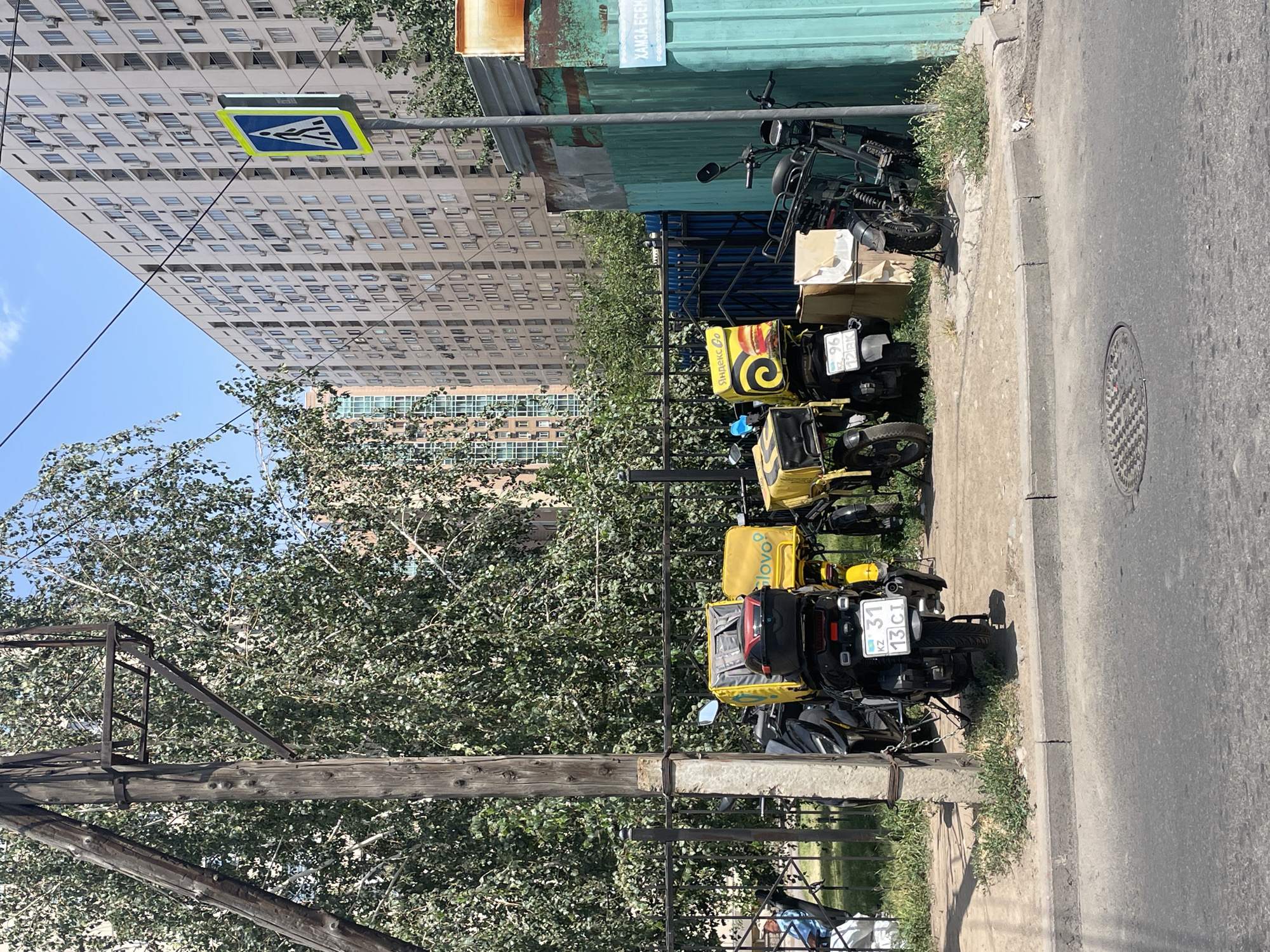 A daytime, eye-level, full outdoor shot captures a street scene in Almaty, Kazakhstan, dominated by several delivery motorcycles parked along a sidewalk. The motorcycles, equipped with large yellow cargo boxes branded with logos such as "Glovo" and "Yandex Go," are positioned near a black metal fence. Behind the fence, lush green trees provide a backdrop. In the distance, tall, modern apartment buildings with numerous windows and air conditioning units rise against a clear blue sky. A pedestrian crossing sign, a yellow triangle with a blue border and a white pedestrian icon, is visible on a pole to the right of the apartment buildings. A turquoise metal fence with rusty patches and a white sign with Cyrillic text is also present on the right side of the image. The foreground shows a paved road with a manhole cover. The scene suggests a typical urban environment in Almaty, likely a busy area with food delivery services operating.