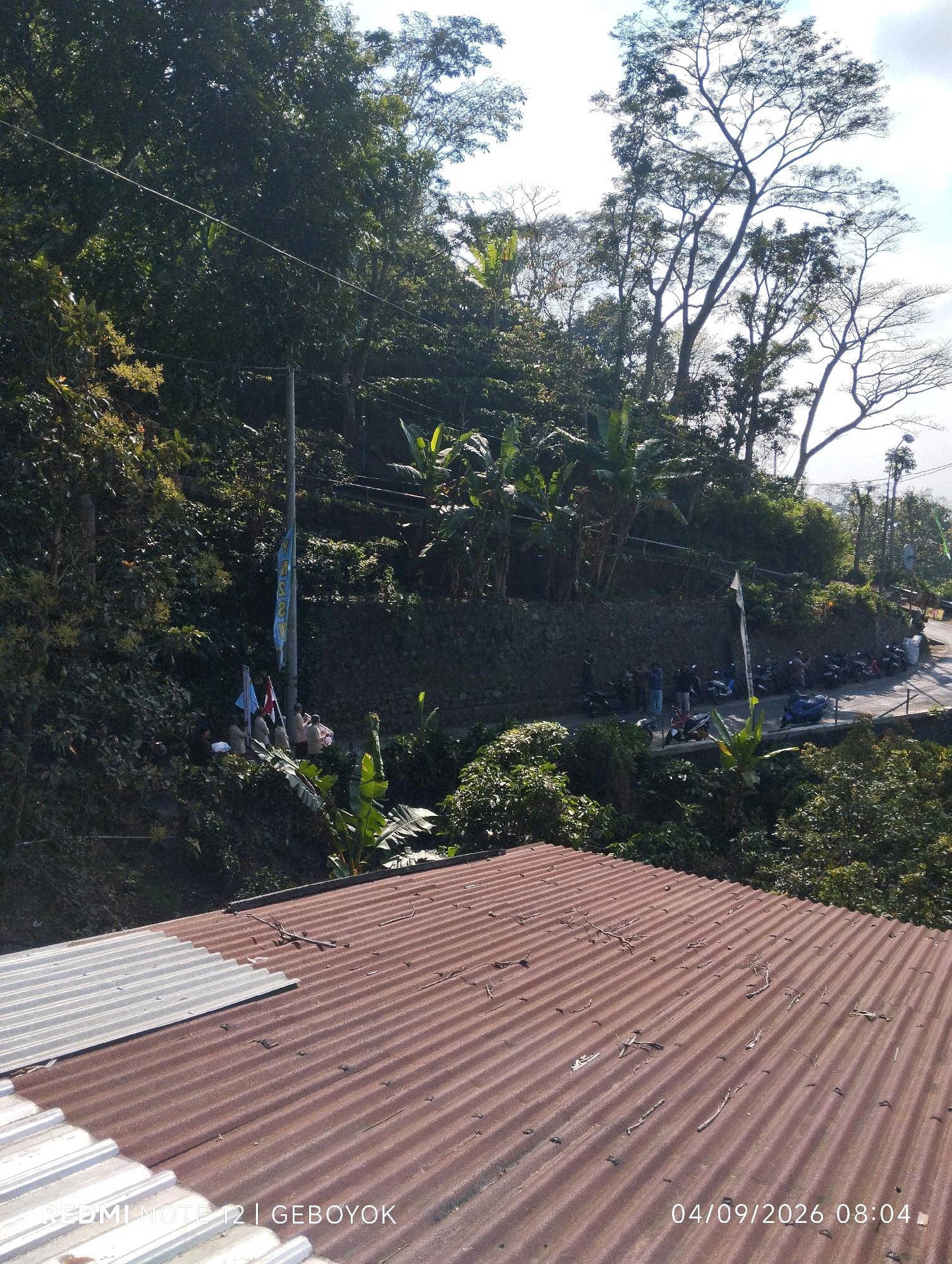 The image depicts a sunlit scene with a strong upward angle, focusing on a steep, tree-covered hillside and a portion of a corrugated metal roof in the foreground. Several motorcycles are parked along a curved road that winds up the hillside. A group of people, some appearing to be wearing uniforms, are gathered near flags on a pole. The environment is lush and green, indicative of a tropical or subtropical climate. The sky is visible in the upper right, with some cloud cover. The overall impression is of an outdoor gathering or event taking place on a scenic, elevated location. The timestamp in the lower right corner indicates the date and time as 04/09/2026 08:04.