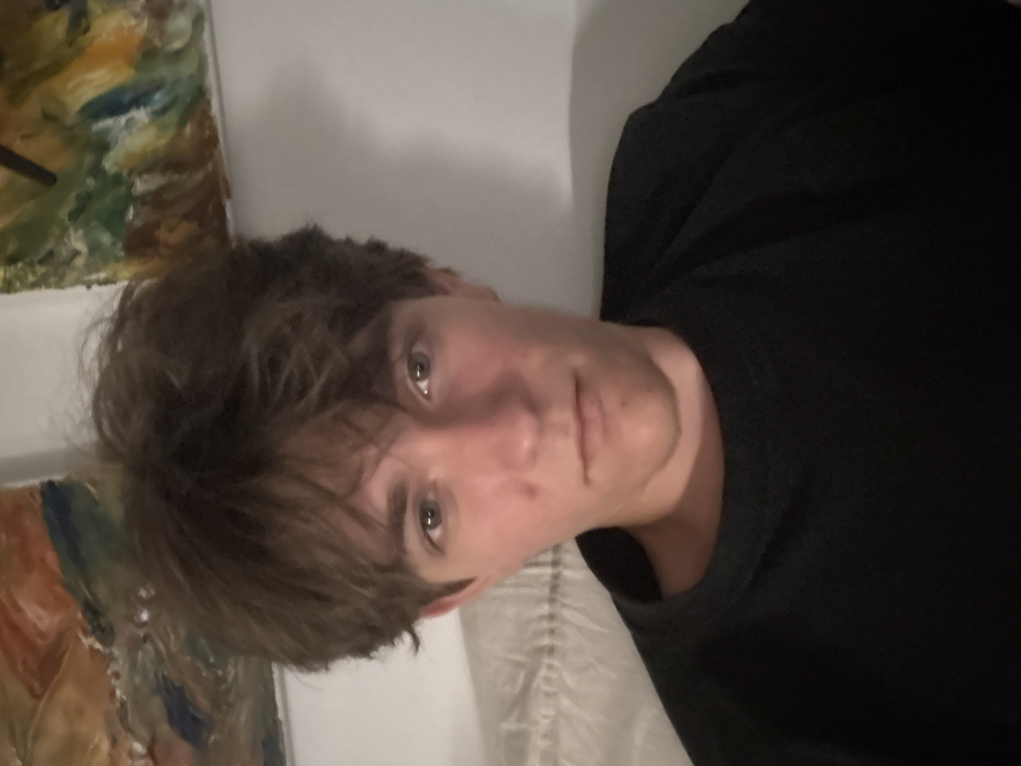 A young person with brown, messy hair is looking directly at the camera. They are wearing a black crew neck shirt and are positioned in front of a wall with abstract paintings. To the left, a white pillow with textured lines is visible. The lighting suggests an indoor environment, possibly a bedroom. There is no information in the image to suggest a location in Cupra Marittima, Italy.
