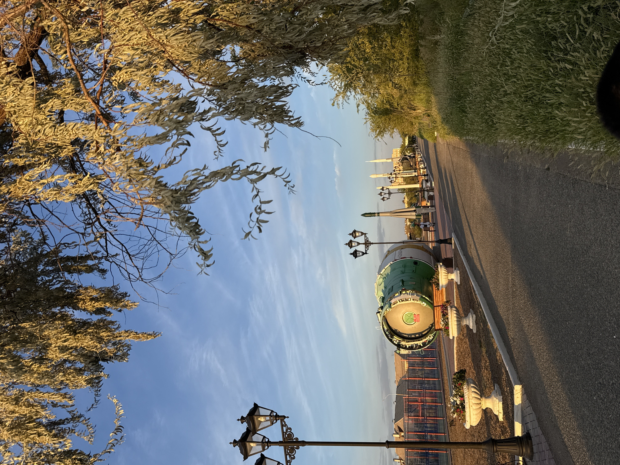 The image depicts an outdoor scene in Baikonur, Kazakhstan, likely in the evening given the golden light. The foreground features a paved walkway and a grassy area. Several elements of interest are visible: decorative lampposts with multiple lights are placed along the walkway. A large, green cylindrical object, possibly a piece of space equipment or a monument, is situated to the left of the walkway, adorned with white decorative urns filled with flowers in front of it. Further down the walkway, another lamppost and what appears to be a rocket are visible, along with several tall, slender structures that resemble minarets or monuments. In the background, buildings and a fence can be seen. A large tree with elongated, golden-hued leaves hangs over the scene from the top left, casting shadows. The sky is blue with wispy clouds. There are a few individuals visible in the distance, walking along the path or sitting on benches. The overall impression is a public space with an exhibition or memorial related to space exploration.