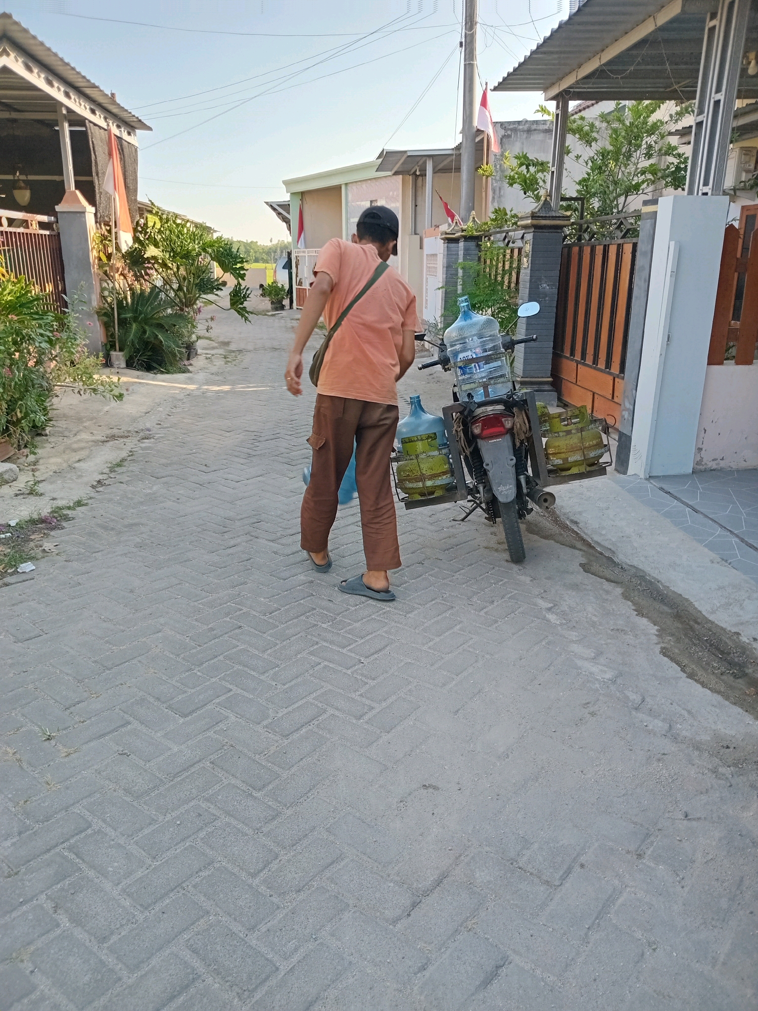 In Mojorayung, Indonesia, a person wearing an orange shirt and brown pants is walking away from the camera. They are carrying a bag over their shoulder and appear to be on a paved walkway. To their right is a motorcycle loaded with large water bottles and gas canisters. The motorcycle is parked next to a fence and a house. The environment suggests a residential area with houses and some vegetation. The sky is clear and bright, indicating it is daytime.