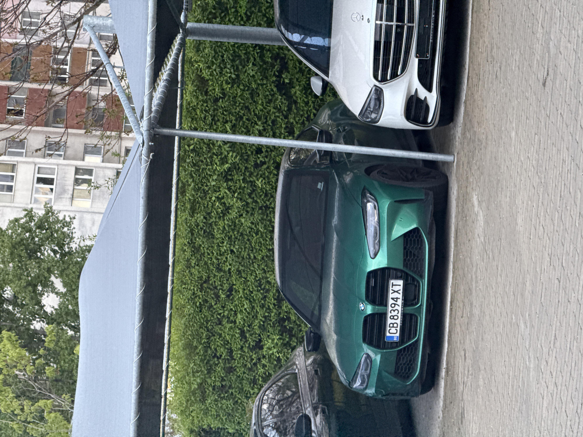 The image shows a green BMW M3 with Bulgarian license plate "CB 8394 XT" parked next to a white Mercedes-Benz. They are parked under a metal-framed shade structure, possibly in a parking lot. A hedge forms a backdrop to the cars. In the background, a multi-story building is visible with multiple windows, suggesting an urban setting. This setting is located in Sofia, Bulgaria, as indicated by the license plate prefix "CB," which is specific to Sofia.