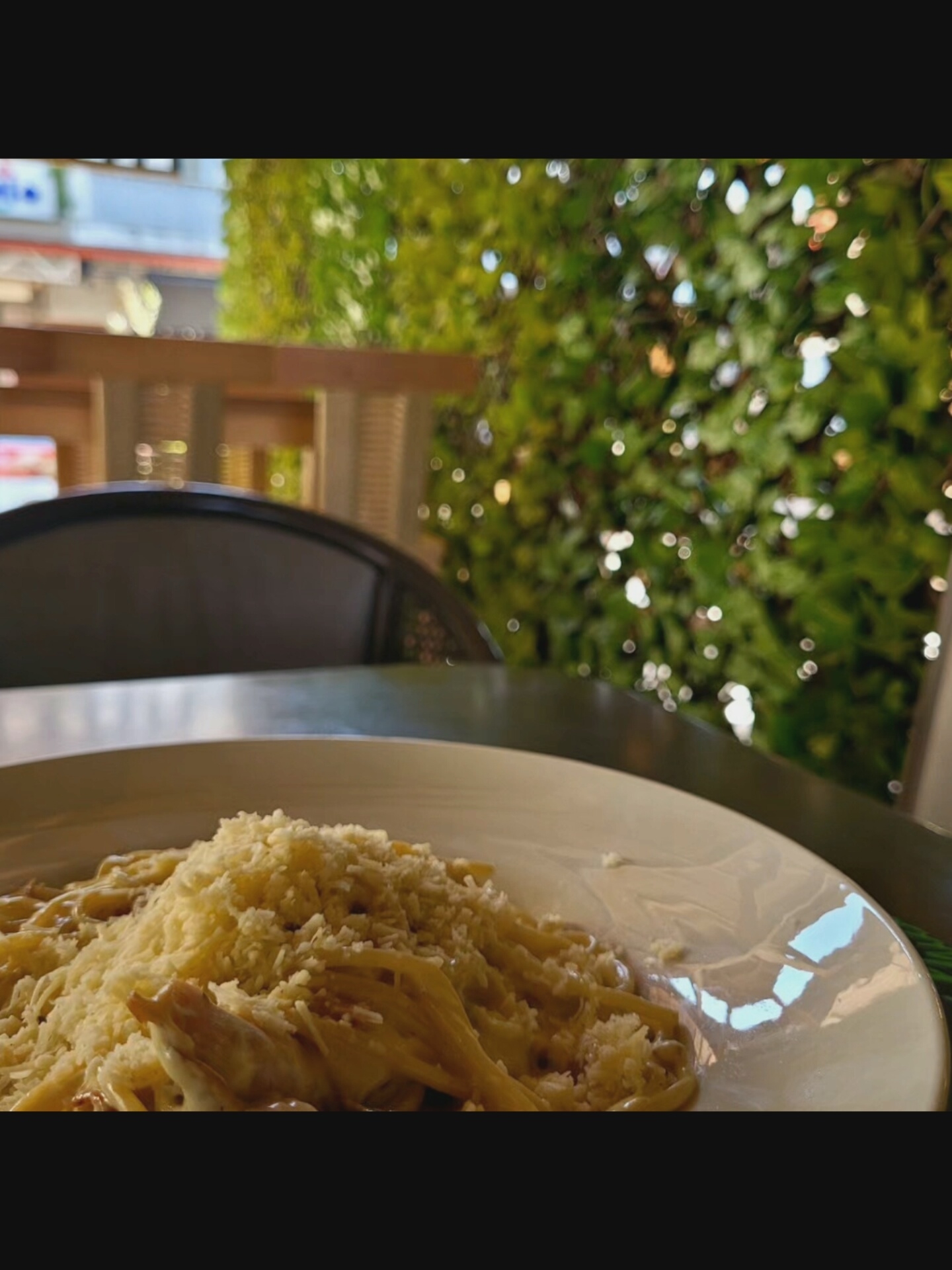 A close-up shot features a plate of pasta, generously topped with shredded cheese. The pasta appears creamy and saucy, with some pieces of meat visible beneath the cheese. The dish is presented on a white, slightly reflective plate. In the background, out of focus, is a vibrant green, leafy wall adorned with small, bokeh lights that create a festive or magical atmosphere. A dark-colored chair is partially visible to the left of the pasta plate. The setting appears to be outdoors, possibly a restaurant patio, with hints of wooden structures and building facades in the blurred distance, suggesting an urban or semi-urban environment in Bielawska Wieś, Poland. The overall mood is inviting and suggests a culinary experience.
