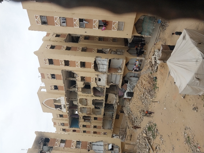 This image captures a scene of severe structural damage to multi-story residential buildings in Khan Yunis, Palestinian Territory, under an overcast sky. The central structure, a multi-story building constructed of beige-colored concrete with arched architectural details, shows extensive damage, with several floors having external walls completely collapsed or blown out, exposing internal rooms and structural elements. The building to its immediate left also exhibits significant damage, including a blackened, possibly charred, upper section.
The ground in the foreground and midground is a vast expanse of sandy earth, heavily littered with concrete rubble, rebar, shattered debris, and piles of broken building materials. In the immediate foreground, a large, light-colored canvas tent is pitched. Several individuals are present: one person walks across the sandy ground in the mid-left, while two other individuals, one appearing to be a child, push a two-wheeled cart through the rubble-strewn area. Another person stands near the tent in the lower right. At the base of the damaged buildings, multiple makeshift shelters or storage areas, constructed from tarpaulins, corrugated metal, and fabric, are visible. Laundry is seen hanging to dry from several windows of the damaged buildings and near the improvised ground-level structures.