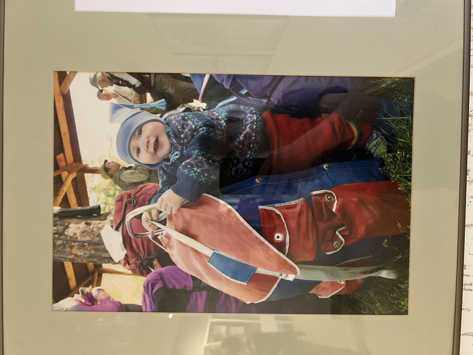 An image displays a smiling baby, appearing to be an infant or young toddler, situated outdoors. The baby is positioned centrally, looking directly at the camera with a wide, joyful smile, revealing a few teeth. They are wearing a light blue knitted beanie with a tassel, a dark blue sweater with a colorful pattern (possibly fair isle style), and red pants.
The baby's hands are holding onto a light red fabric item that resembles a climbing rope bag or a gear sling, with a white strap and a blue patch, and metal carabiners visible near its top edge. This item rests on a larger blue fabric surface, possibly a mat or another piece of outdoor gear. Behind the baby, green grass is visible to the right.
In the blurred background, several adults are partially visible, one wearing a hat and another with a backpack. A wooden structure, possibly part of a building or shelter, is also present, suggesting a rustic or natural setting. Other outdoor gear, including red and purple backpacks, can be seen in the background. The overall scene conveys a cheerful outdoor activity, likely in a natural or resort-like environment such as Sanatoriya Yumatovo Imeni 15-Letiya Bassr, Russia, during daytime under soft, natural light. No discernible text is visible within the image.