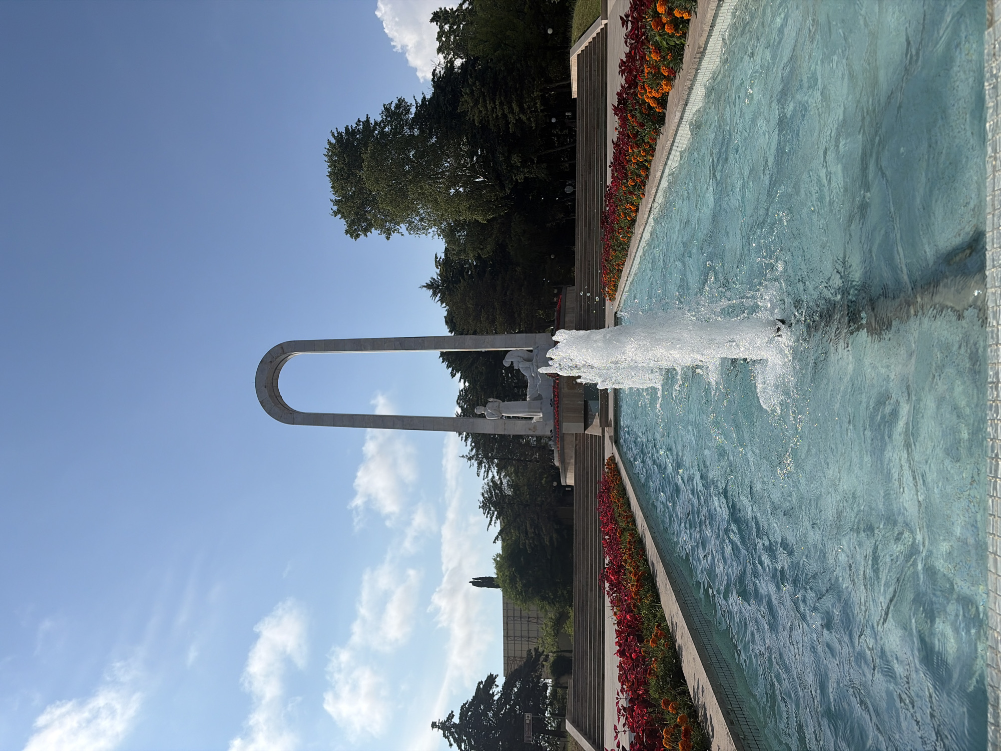 This media file depicts an outdoor scene featuring a large fountain with a powerful jet of water spraying into the air. The fountain is lined with a strip of colorful red and orange flowers, and beyond that, a set of wide concrete stairs ascends. At the top of the stairs, a monument with statues of figures is visible. In the background, lush green trees create a dense canopy, suggesting a park or garden setting. The sky is a clear, bright blue with a few fluffy white clouds, indicating pleasant weather during the daytime. The composition is a bit unusual due to the rotation of the image, with the fountain and stairs running vertically on the right side of the frame. The overall impression is one of a serene and well-maintained public space. No text is visible in the image.