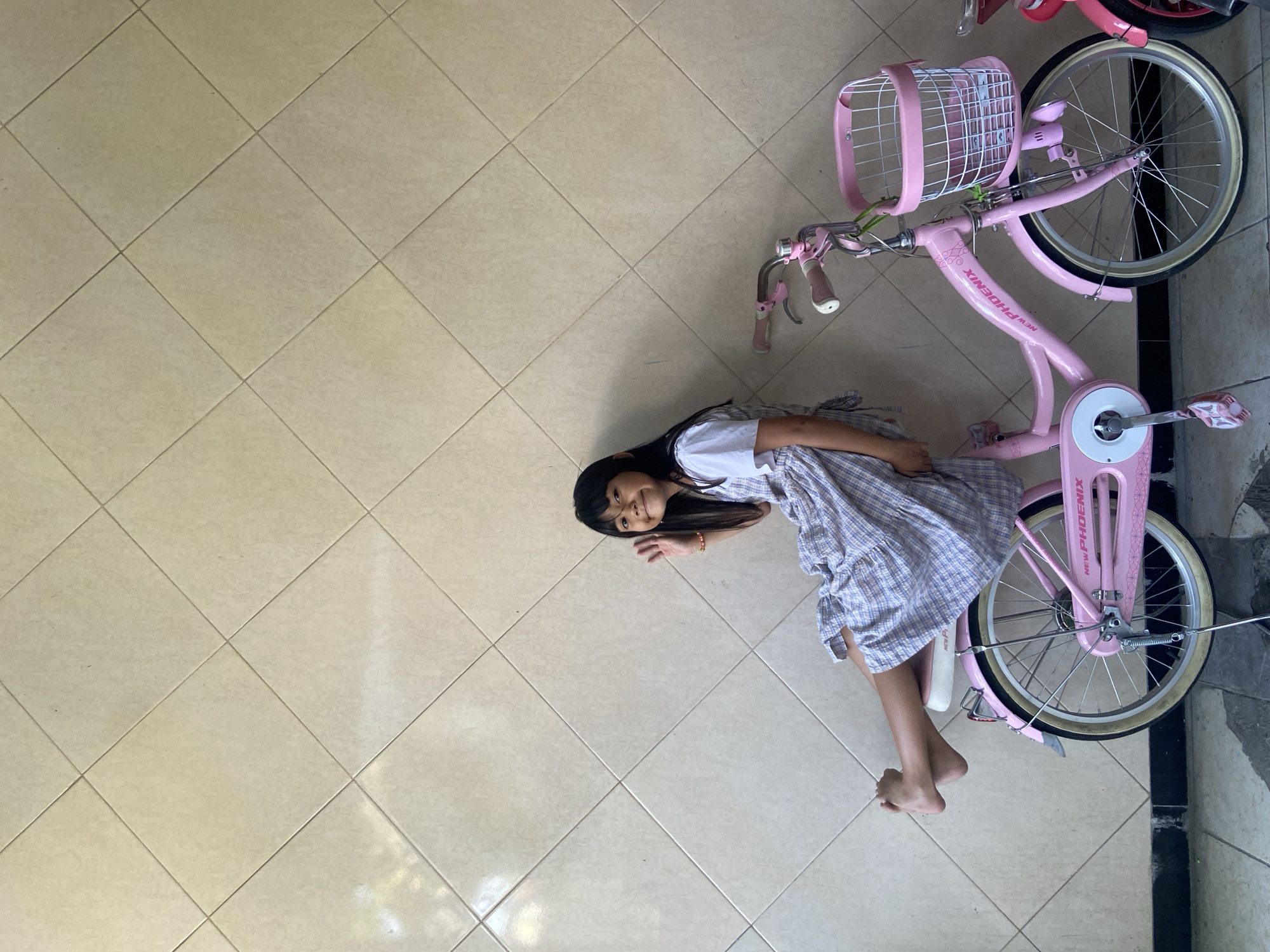 A young girl with dark hair, wearing a light blue and white plaid dress over a white t-shirt, sits on the seat of a pink bicycle. Her left leg is extended and resting on the rear wheel fender, with her bare feet pointing upwards. Her right leg is bent, with her foot resting on the ground near the bicycle's pedal. She is smiling and waving at the camera with her right hand. The bicycle is pink with a white seat, a white basket on the handlebars, and a silver kickstand. The word "PHOENIX" is visible on the bicycle frame. The background consists of a wall tiled with light beige square tiles arranged diagonally, and a dark border along the bottom. The scene is outdoors, possibly on a street or pathway in Gelam, Indonesia. Another pink bicycle is partially visible to the right.