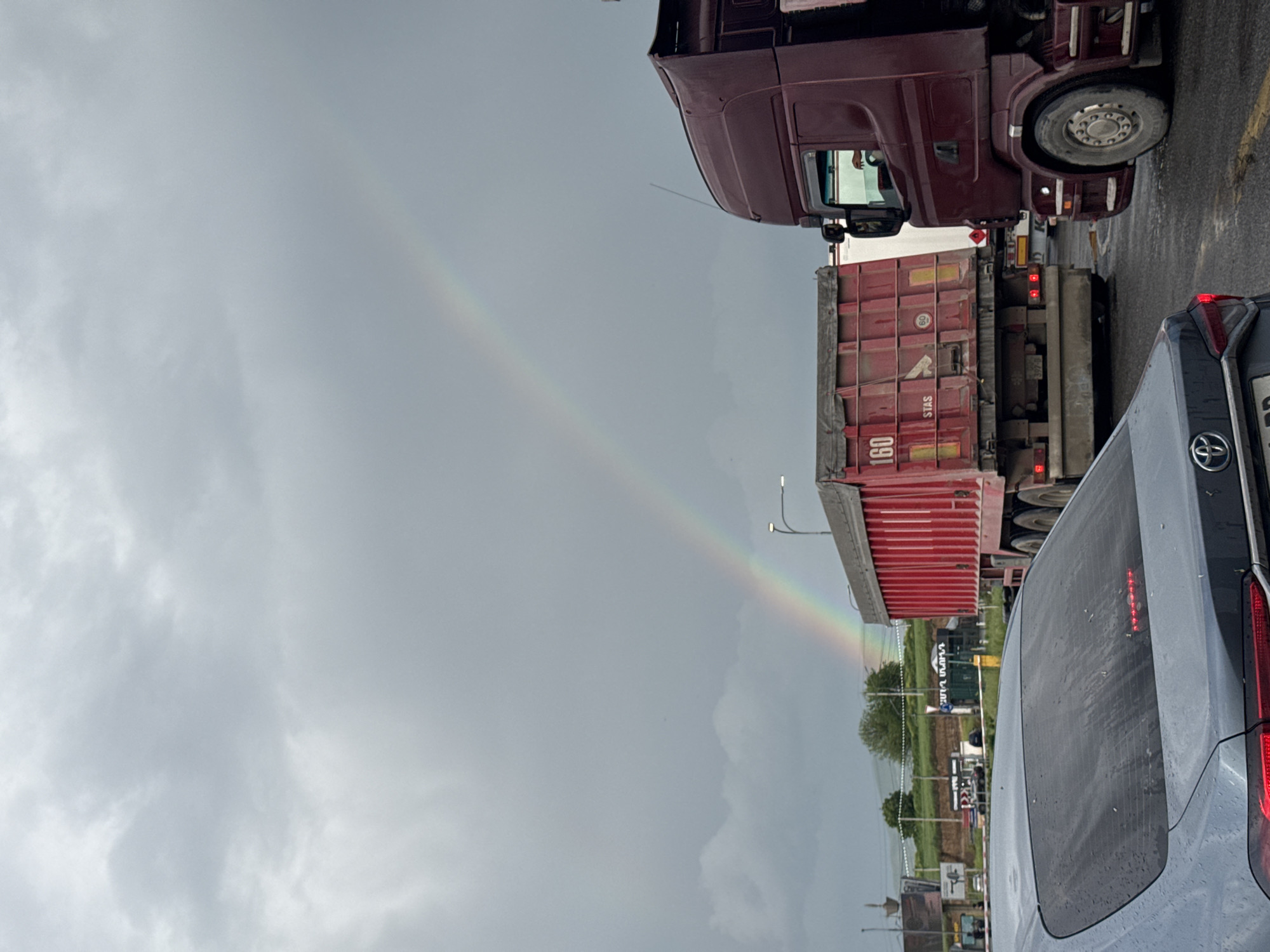 The image shows a rainbow stretching across a gray, overcast sky in Bagratashen, Armenia. Below the rainbow, there is a road with cars and trucks, including a red semi-truck with a shipping container and a gray car with a Toyota logo. The ground features greenery and various structures, suggesting an industrial area or border crossing. The picture is taken from behind the gray car.