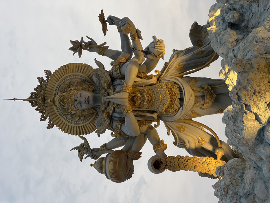 A monumental, multi-armed statue, predominantly pale grey with extensive golden detailing, stands against a light, overcast sky in Kerobokan Kelod, Indonesia. The central figure, representing a deity, features a serene facial expression with red lips, adorned with a golden crown, ornate jewelry, and a prominent golden sun-ray halo behind the head. Two central hands are pressed together in *Anjali Mudra*. A total of ten arms extend from the torso. On the left side, an upper hand cradles a golden, ornate pot, while a lower hand pours a cascade of golden circular objects, resembling coins, downwards towards the base. On the right side, an upper hand gently holds a golden lotus flower bud, and a lower hand holds a flat, open lotus bloom or offering. Other hands are posed or hold smaller, less distinct items. The statue wears elaborate golden garments and stands upon a textured, rocky-like base. The base and lower portions of the statue are illuminated by warm, artificial lights, creating strong contrasts and highlights on the golden elements.