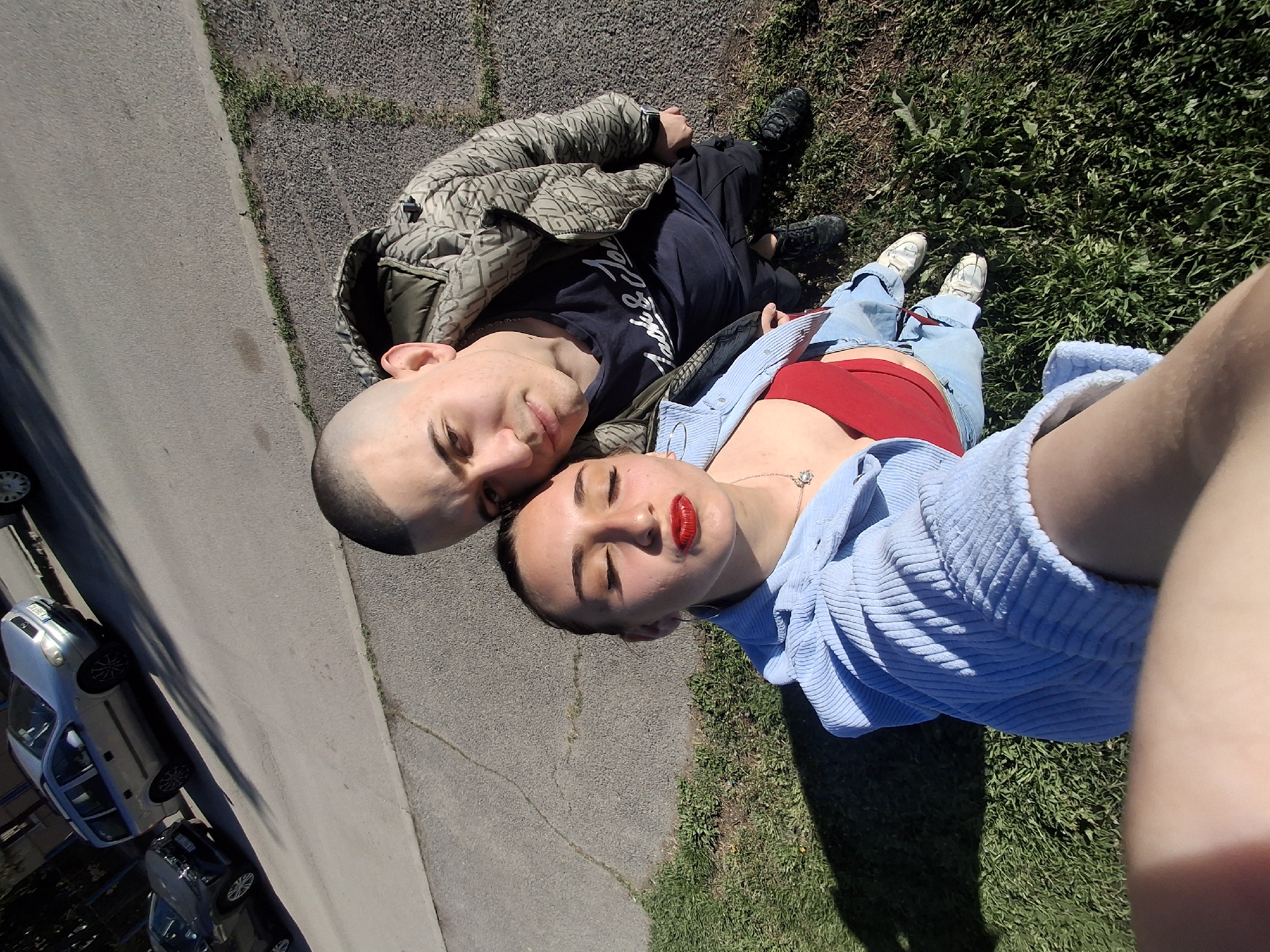 Two light-skinned individuals lie on the ground, positioned on a grassy patch adjacent to a paved road in Sofia, Bulgaria, as evidenced by the license plates of the vehicles parked on the roadside. The male, with a shaved head, wears a dark t-shirt and a patterned coat, and is lying on an area of dark gravelly material. The female, with bright red lipstick, wears a red top and light blue denim jacket, lying on the grass. The camera angle is angled downward as though the female subject is taking a selfie. Sunlight casts shadows on the ground.