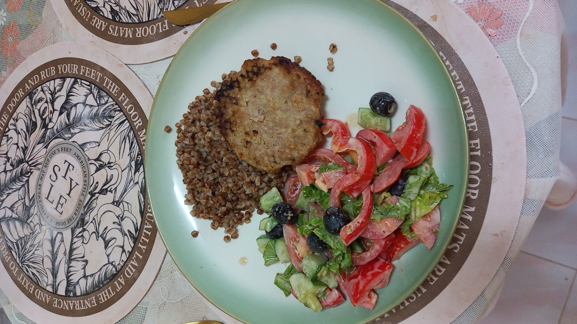 An overhead medium shot captures a plate of food presented on a table, possibly in a restaurant setting in Koh Phangan, Thailand. The plate, light green with a white rim, holds a meal consisting of cooked buckwheat, a pan-fried cutlet, and a salad. The salad is a colorful mix of chopped lettuce, sliced tomatoes, cucumbers, and black olives.
The table is adorned with two circular placemats, one partially visible on the left and the other more prominently displayed on the top of the image. These placemats feature an intricate, dark-colored design with feathered motifs and text that appears to be in English, though not fully decipherable from the image. The text around the edge of the top placemat reads, "QUALLY LAID AT THE ENTRANCE AND EXIT TO PRE... THE DOOR AND RUB YOUR FEET THE FLOOR MAT..." and "...FLOOR MATS ARE USUA".
A patterned tablecloth, predominantly white with subtle floral accents in pink and beige, lies beneath the placemats and plate. The lighting suggests an indoor environment, possibly daytime given the overall brightness. There are no people visible in the frame, and no specific indication of the exact location within Koh Phangan, other than the general context of a food presentation.