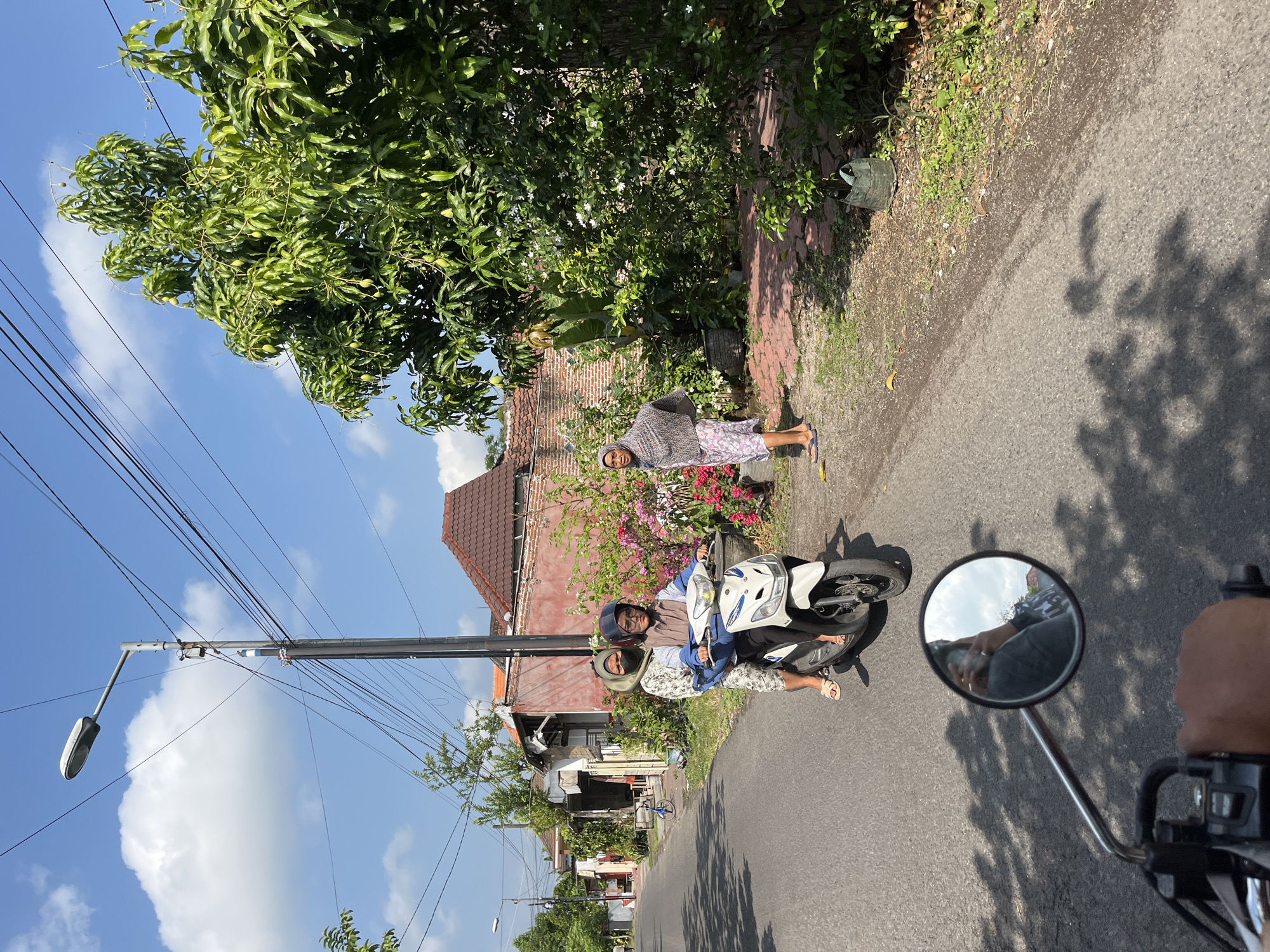 In Jati Alun Alun, Indonesia, on a sunny day, two women ride a white scooter down an asphalt road. The woman in the front wears a light brown hijab and a blue shirt, while the woman in the back has a light green hijab and a patterned dress. They are both looking forward. To the right of the road, a woman in a patterned scarf and a pink and white skirt stands next to a lush green tree and flowering bushes. The background features a red-tiled roof building and houses lining the street. Overhead, a street lamp stands tall amidst a network of electrical wires against a bright blue sky with scattered clouds. In the foreground, a motorcycle's rearview mirror reflects the driver and the sky.