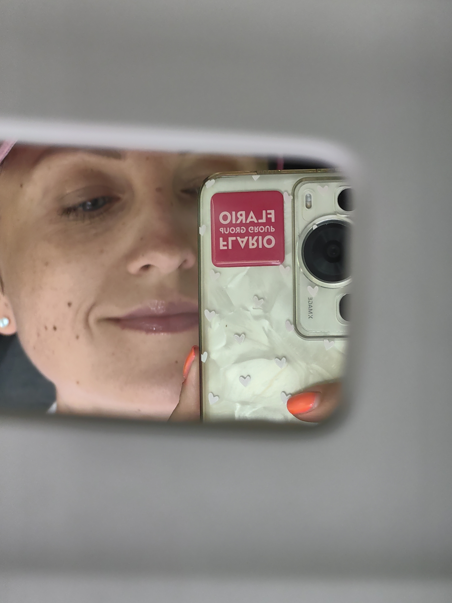 The image shows a reflection in a mirror. A woman's face, including her eye, nose, and mouth, is visible. She is wearing a pink hair covering. Her phone, with a white case decorated with small pink hearts, is reflected in the mirror, and she is holding it with an orange-painted fingernail. The phone has a pink sticker with white text that reads "ELGARIO" in uppercase letters, and below that, "ELGARIO GROUP" in a smaller font, all reversed as it is a reflection. The background of the reflection is a light gray, and the overall scene is lit. The image does not provide information about the city of Хаценчыцкі сельскі Савет, Belarus.