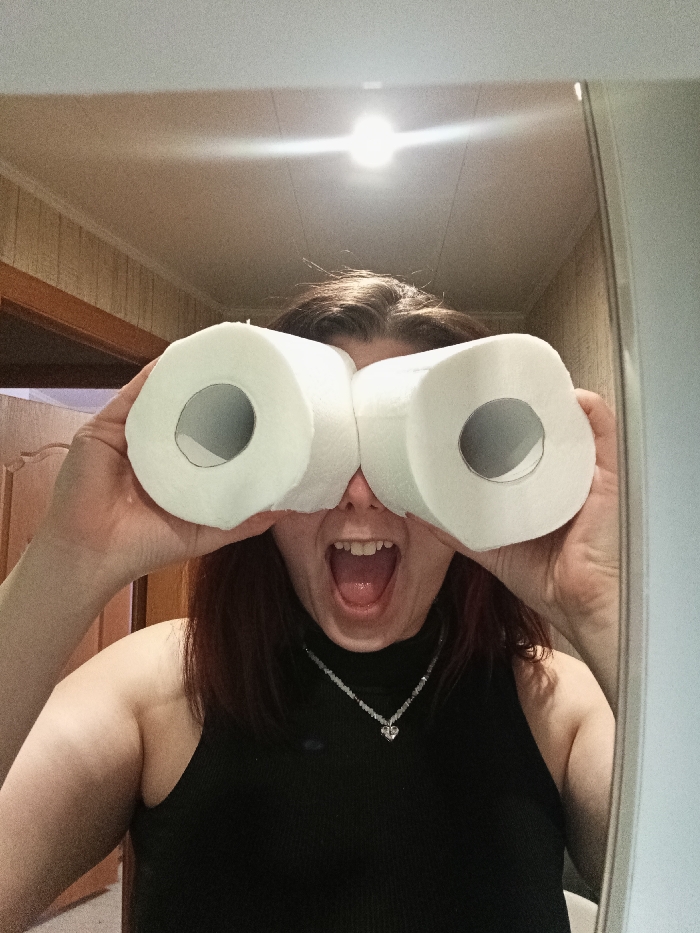 The image shows a person, likely female, from the chest up, engaged in a playful activity indoors. The person has dark brown hair and is holding two rolls of white toilet paper up to their eyes, mimicking binoculars. Their mouth is wide open in an expression that conveys surprise, excitement, or humorous awe, revealing teeth and tongue. Their eyes are completely obscured by the toilet paper rolls.
The person is wearing a black, sleeveless, high-neck top. Around their neck, a delicate necklace with small, light-colored beads and a prominent heart-shaped pendant is visible. Both hands are holding the toilet paper rolls firmly against their face. The cardboard cores of the toilet paper rolls are visible through their centers.
The background suggests an indoor setting, possibly a bathroom or hallway, with light-colored walls. On the left side of the frame, there's a section of what appears to be wood paneling. A bright light fixture is visible at the top center of the image, casting a strong glare or reflection. The composition suggests the photo was taken as a selfie in a mirror, with a faint vertical line on the right side possibly indicating the edge of the mirror. The overall mood of the image is lighthearted and humorous. There is no visible text in the image.